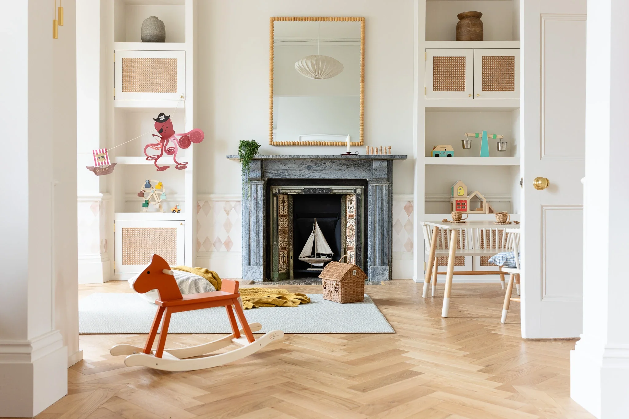 A cozy children's playroom with a decorative fireplace, white built-in shelves filled with toys and decorative items, a mirror framed with natural wood, and a rocking horse in the foreground. The room has light hardwood floors and a mix of modern and playful decor.