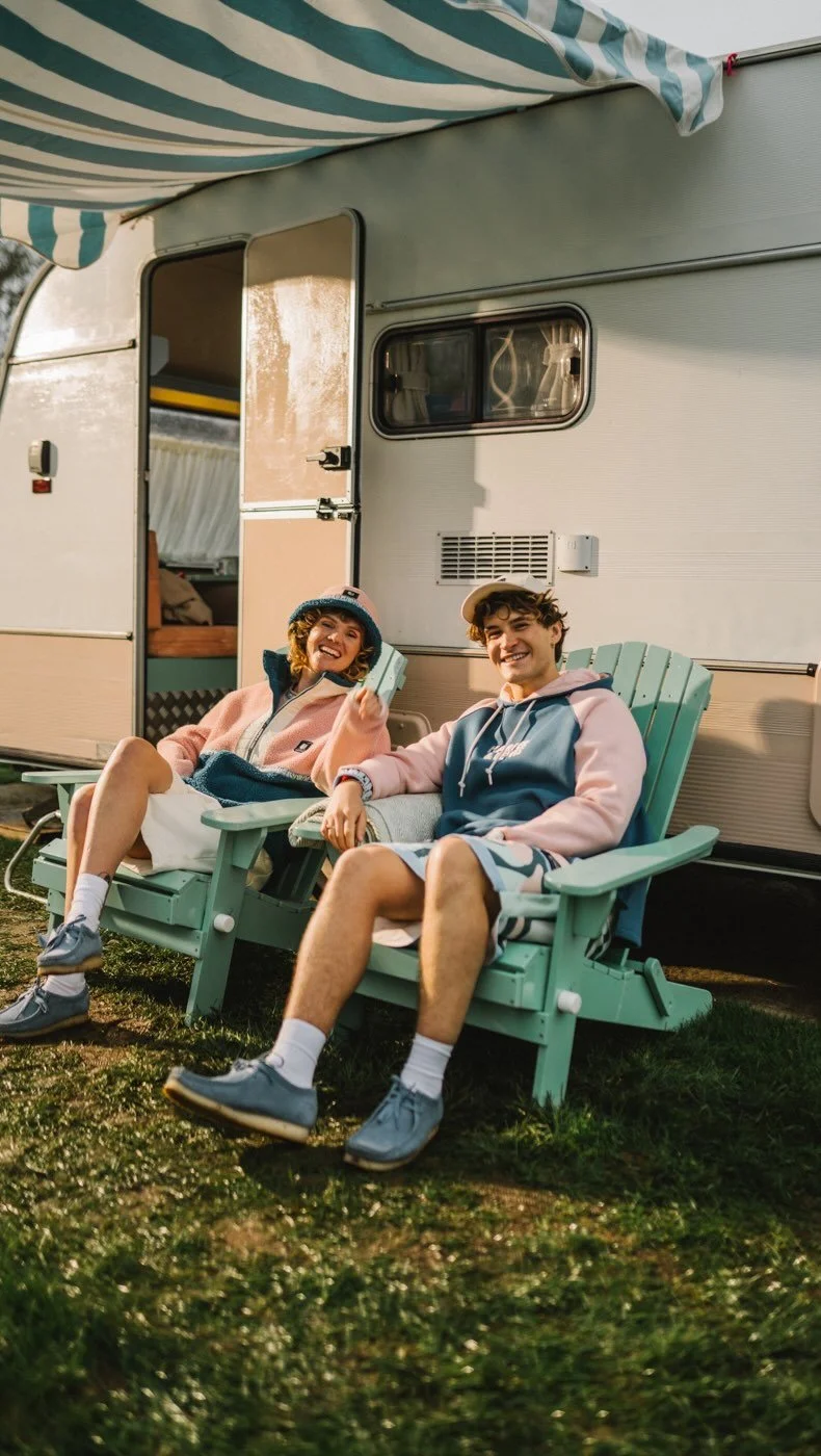 Two young people sitting in green Adirondack chairs outside a camper trailer, smiling and relaxing. The woman is holding a phone, and both are dressed casually in hoodies, shorts, and sneakers.