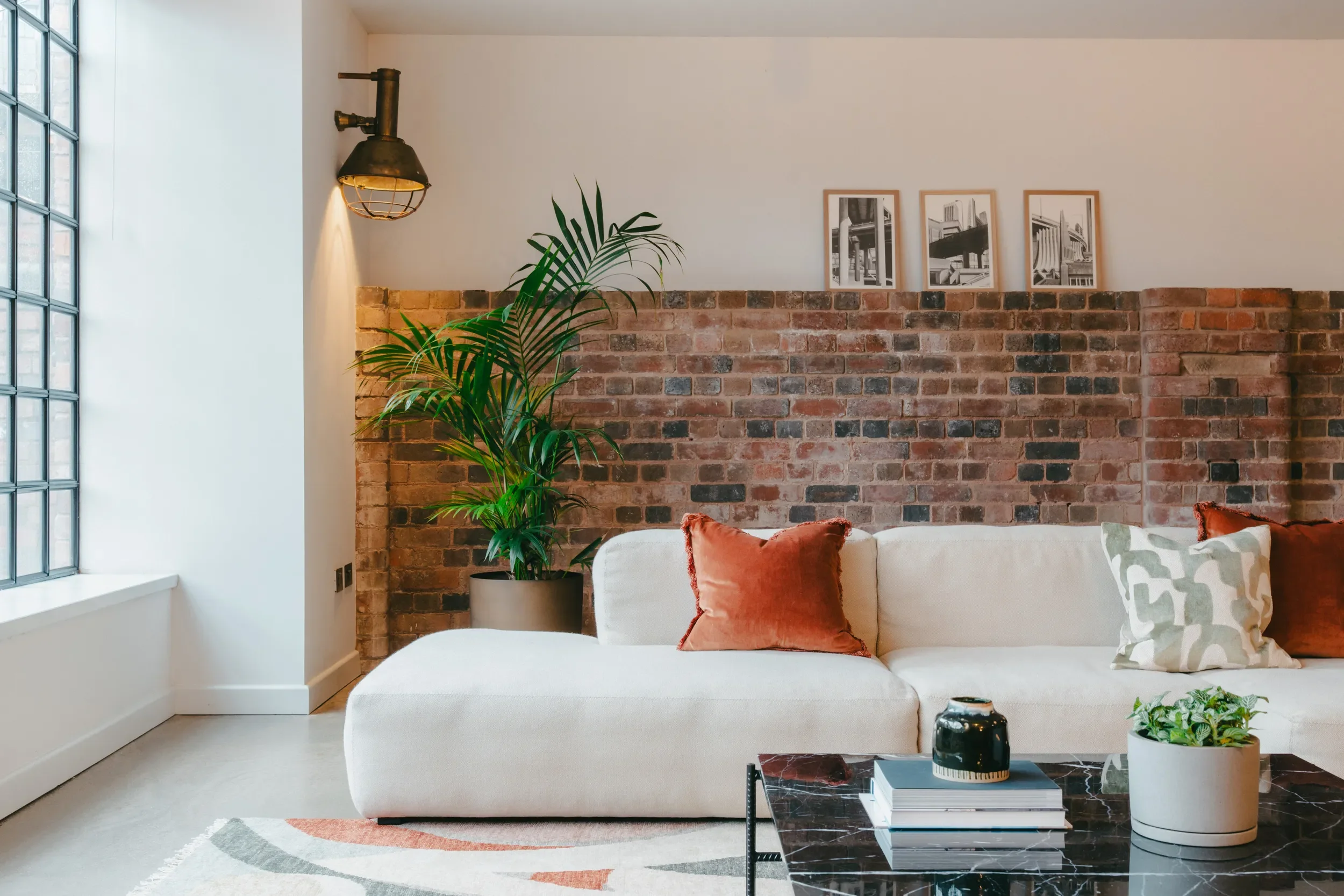 Modern living room with a large beige sofa, rust-colored pillows, a black marble coffee table with books and decorative items, a green plant, a brick accent wall, three framed black-and-white prints, a window with a grid pattern, and an industrial-style wall sconce. Style is minimalistic and contemporary.