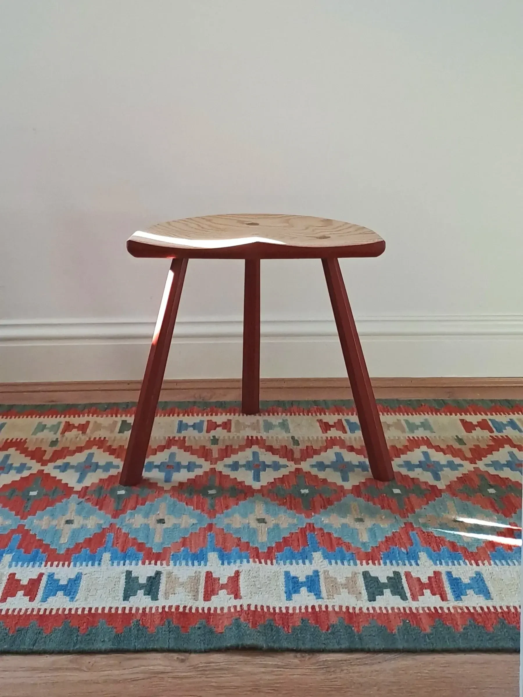 A wooden stool with three legs stands on a colorful, patterned rug in front of a plain white wall.