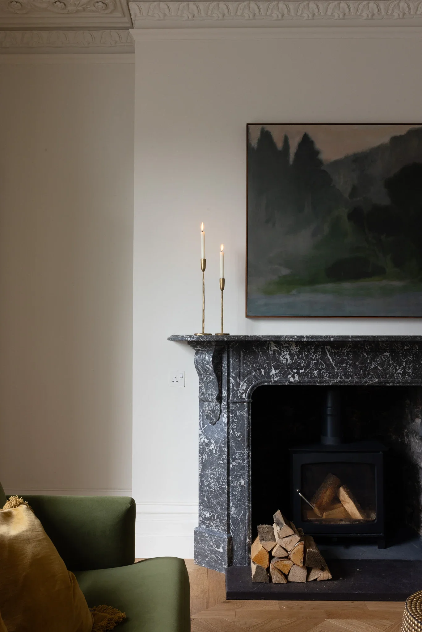 A living room corner featuring a black marble fireplace with a wood-burning stove, stacked firewood underneath, a large landscape painting above, two tall lit white candles on a gold holder on the mantel, and part of a green armchair with a throw pillow visible in the lower left.