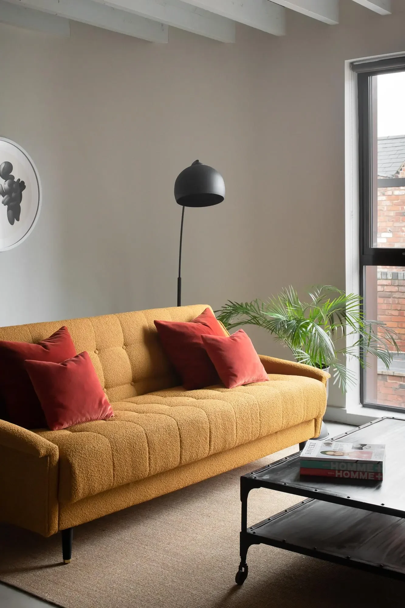 A living room corner with a mustard yellow sofa, red velvet pillows, a black floor lamp, a window with a brick building in view, a potted plant, and a coffee table with magazines.