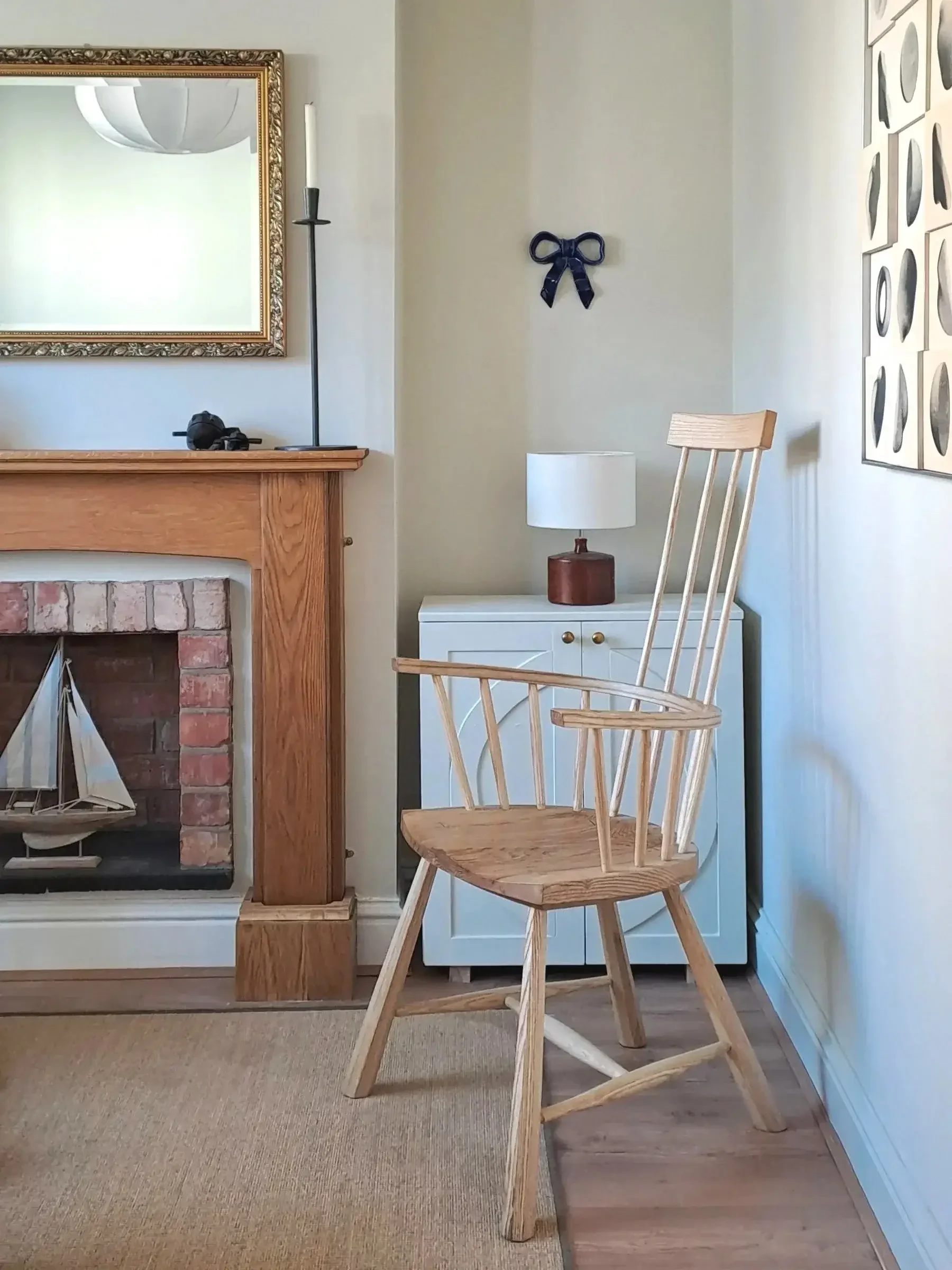 A cozy interior corner with a wooden chair, a white side table with a lamp, a fireplace with brick interior and a wooden mantel, a large mirror with ornate gold frame, a black castle model on the mantel, a black ribbon decoration on the wall, and a series of black and white circular wall art on the right.
