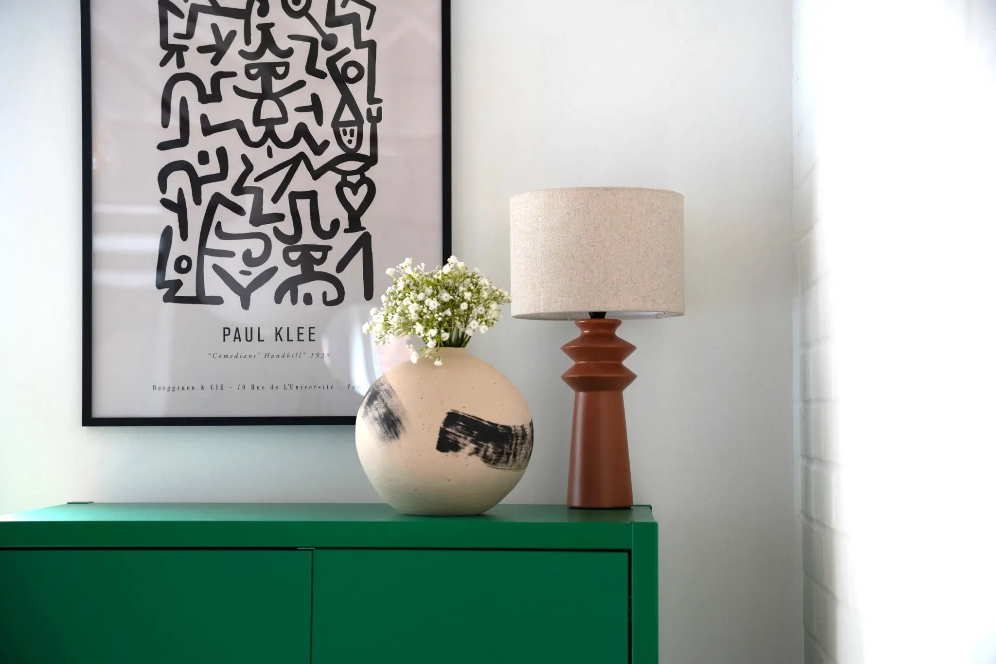 A framed black and white abstract art print on the wall, featuring a face with a smile, and the text 'Paul Klee' and 'Comedians' Handbill' 1938. Below the print is a green cabinet with a beige and black pottery vase holding white flowers, and a tan ceramic table lamp with a textured cylindrical shade on top.