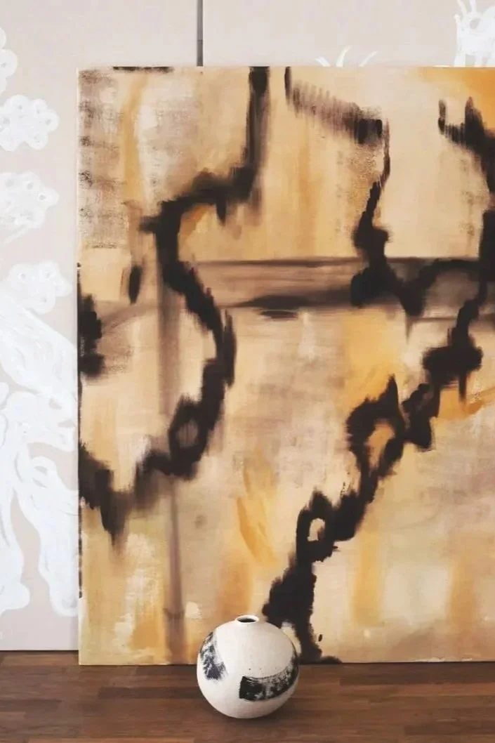 Abstract painting with beige, black, and white brushstrokes leaning against a wall. A round vase with black markings is placed in front of the painting on a wooden floor.