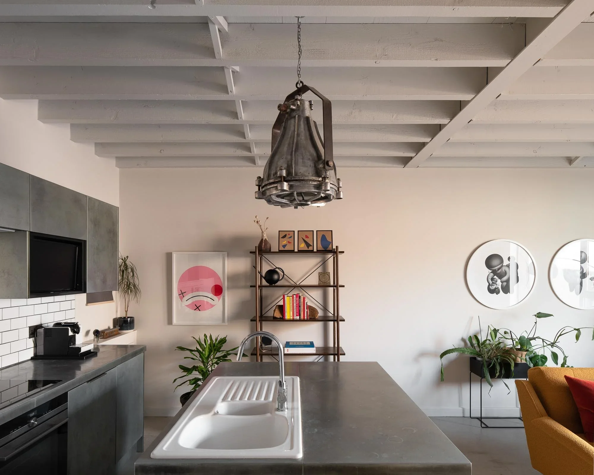 Modern kitchen with gray cabinets, a white subway tile backsplash, a black coffee machine on the counter, and a white sink with a silver faucet in the foreground. A wooden shelf with framed art, books, and decor is on the wall, with various potted plants nearby. A metallic industrial-style chandelier hangs from a white exposed ceiling with wooden beams.
