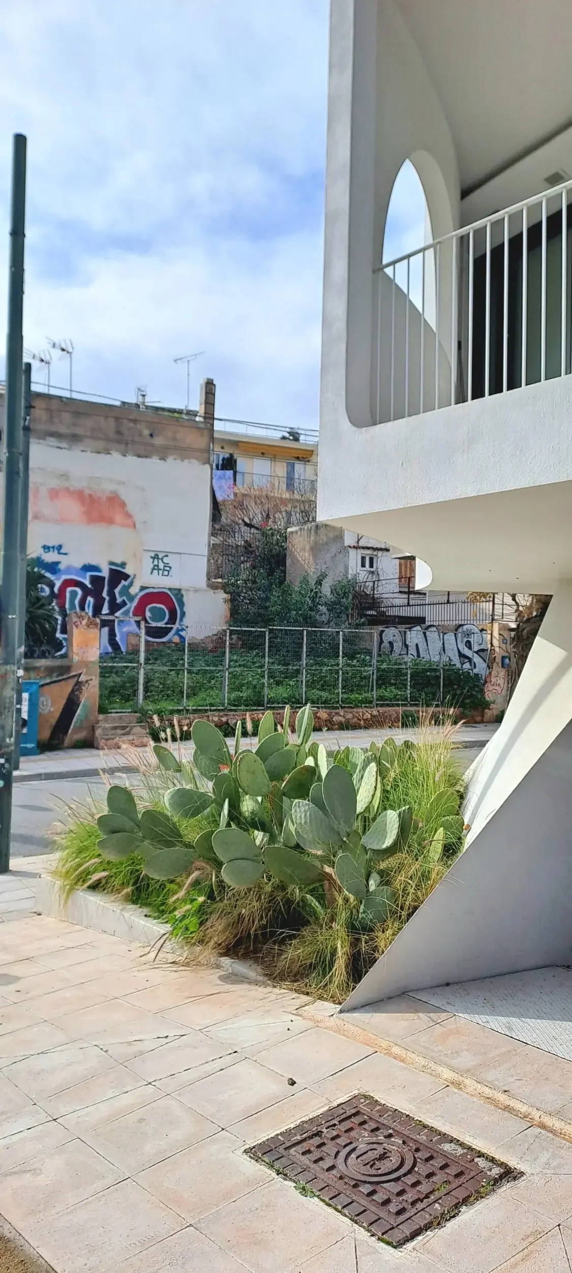 The image shows a small outdoor area with green plants, including cactus, in a plaza. There is a tile floor, a drain, and a white modern building with a balcony. In the background, a street and disturbing graffiti art on a wall are visible under a cloudy sky.