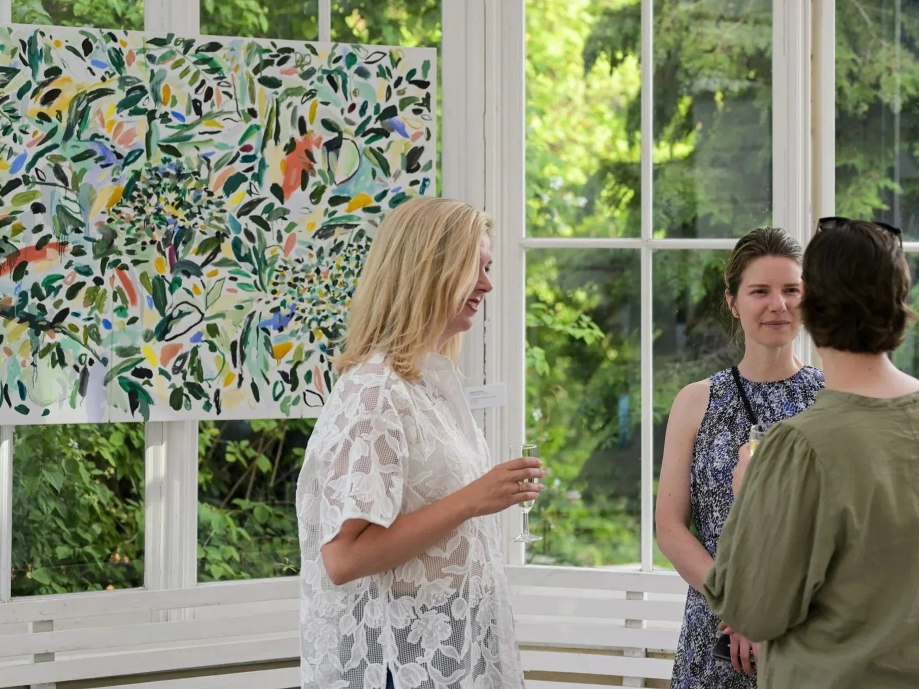 Three women at an art gallery or exhibition, standing near a window with lush green trees outside. One woman is wearing a white lace dress, holding a drink, smiling. Another woman in a sleeveless patterned dress is engaged in a conversation, and the third with glasses and short dark hair is partly visible, facing the second woman.