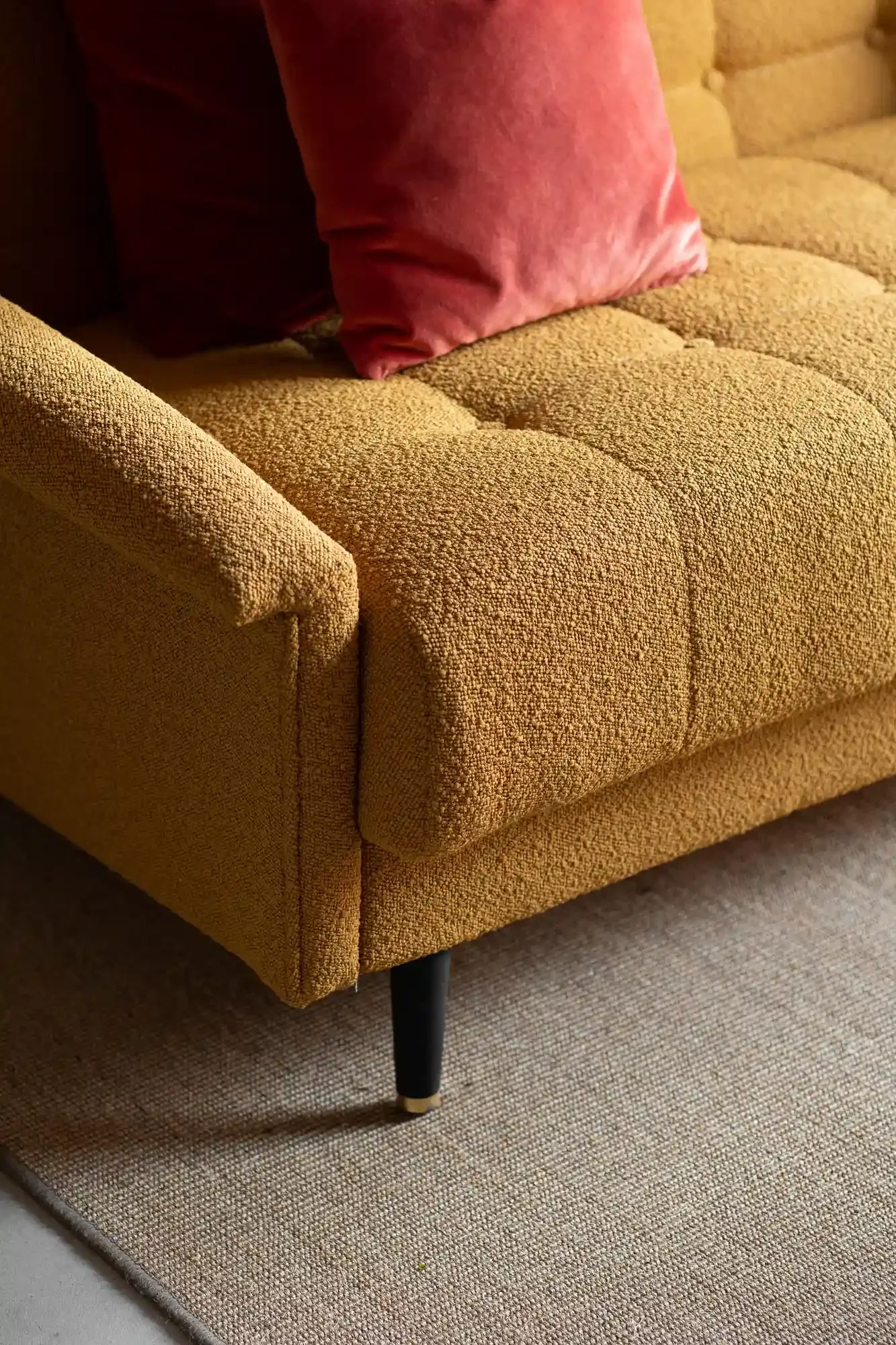 Close-up of a mustard yellow textured sofa with black legs, a pink velvet pillow, and a beige woven rug underneath.