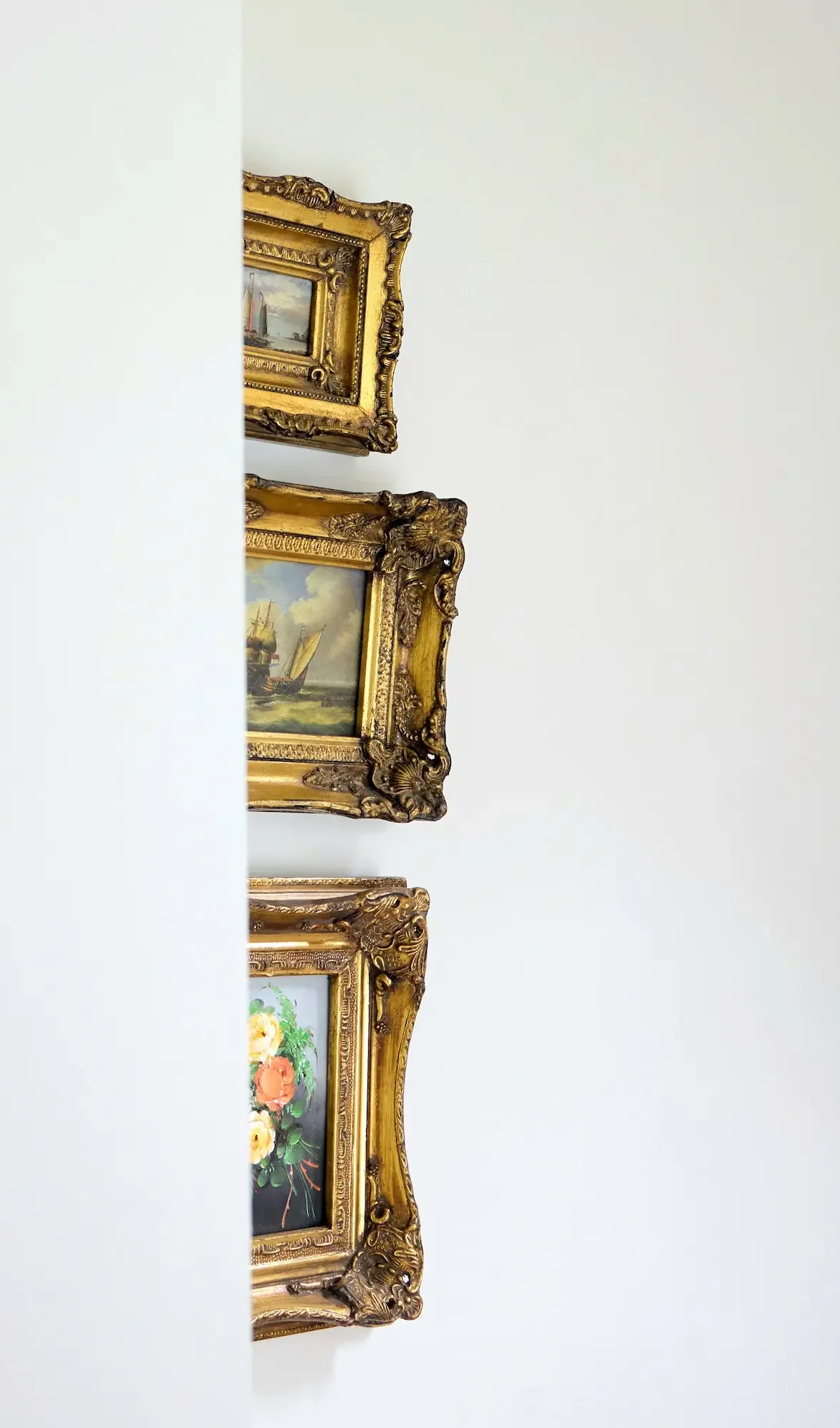 Close-up of ornate gold-framed paintings hanging on a white wall, with several paintings visible, including a ship and a bouquet of flowers.
