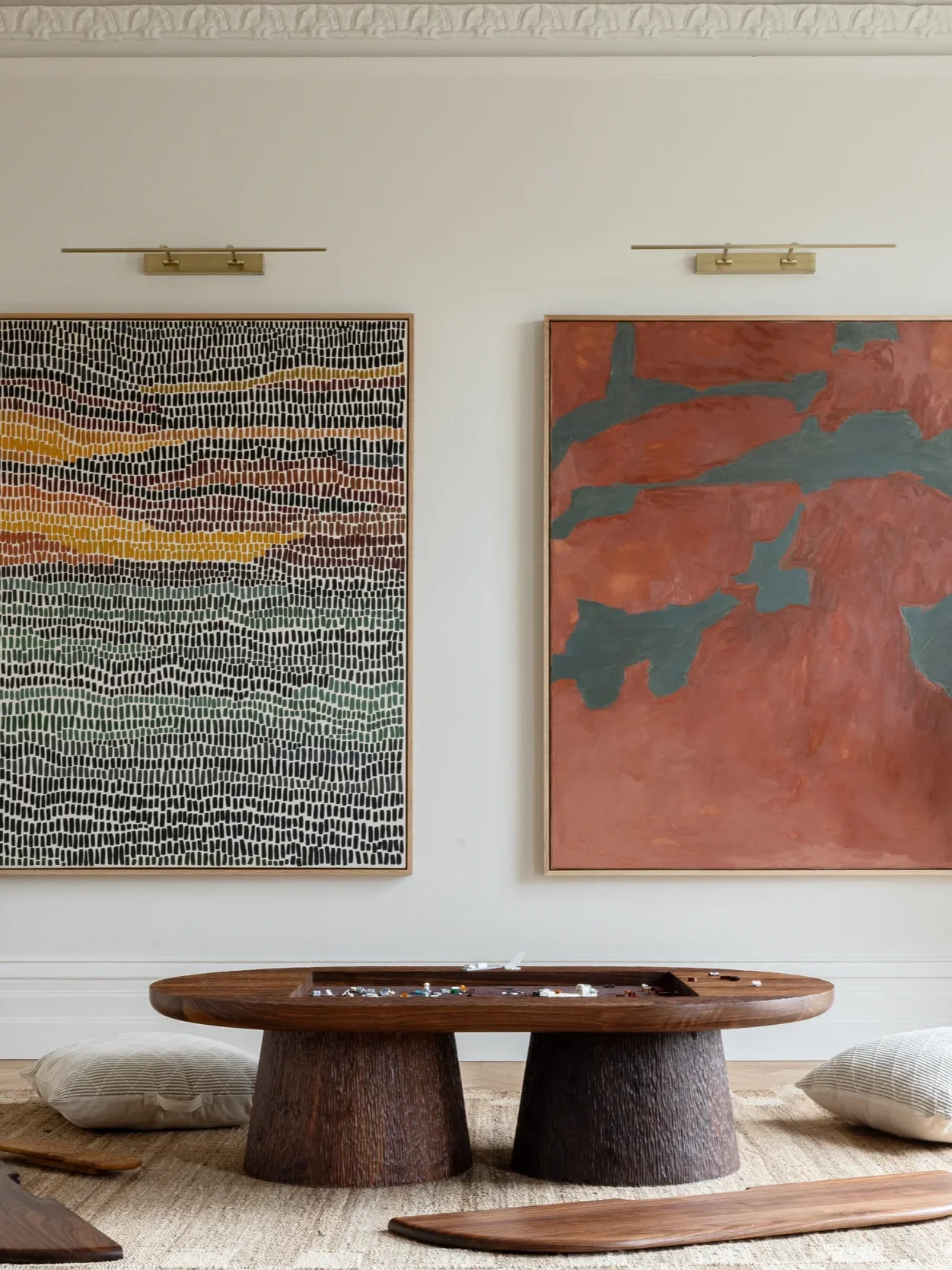 Living room with two large abstract paintings hanging on the wall, a low wooden coffee table with jewelry on it, and two beige pillows on the floor.