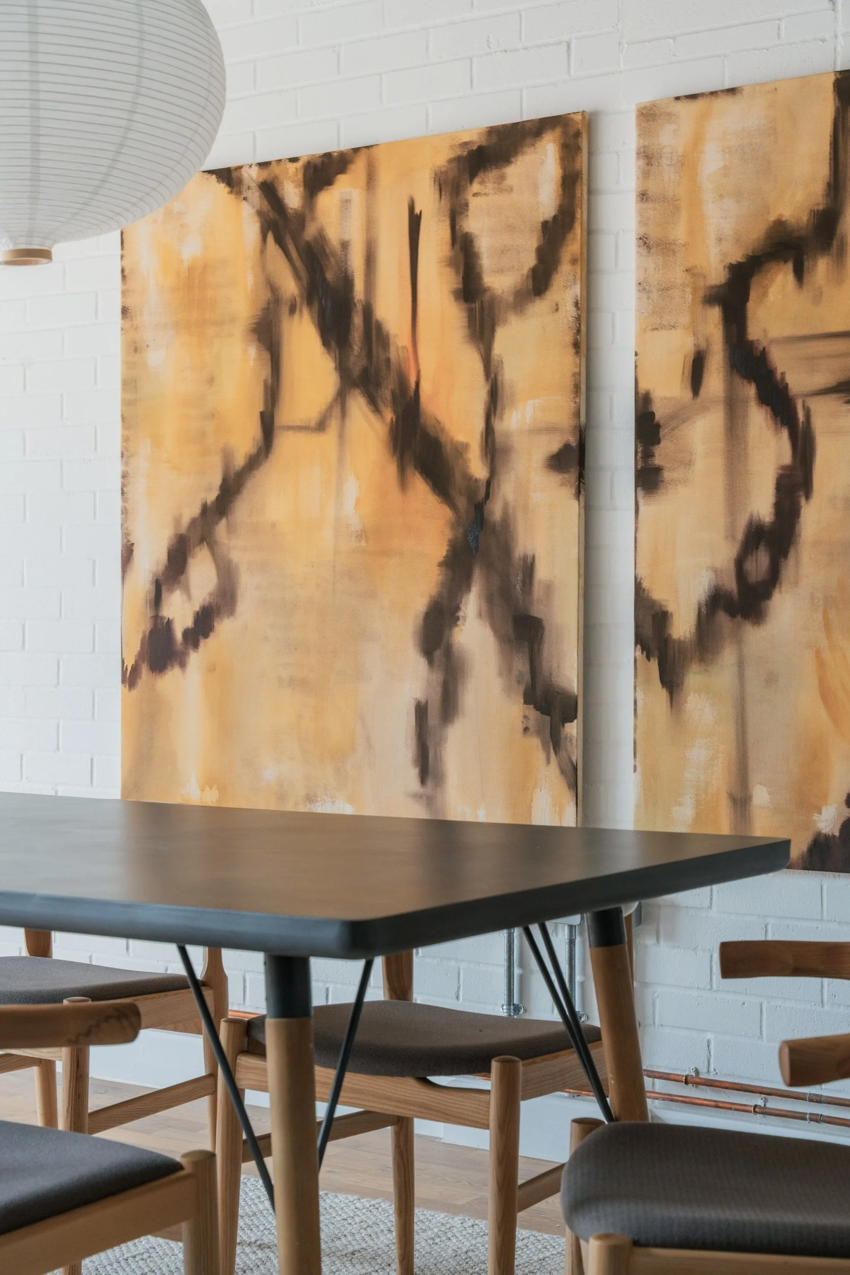 Two large format canvas on a white brick wall. Painted by Aly Grimes, founder and director of San Stae Interior Design and Art Studio. Large paper lampshade in the top left corner.