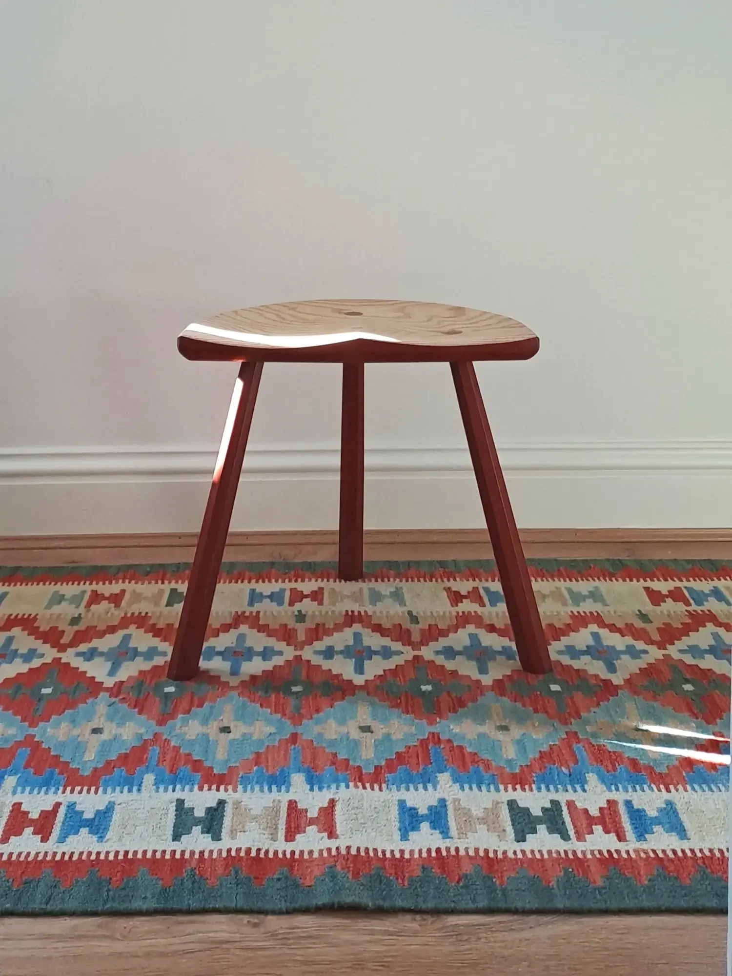 A simple wooden stool with three legs placed on a colorful patterned rug in front of a plain white wall.