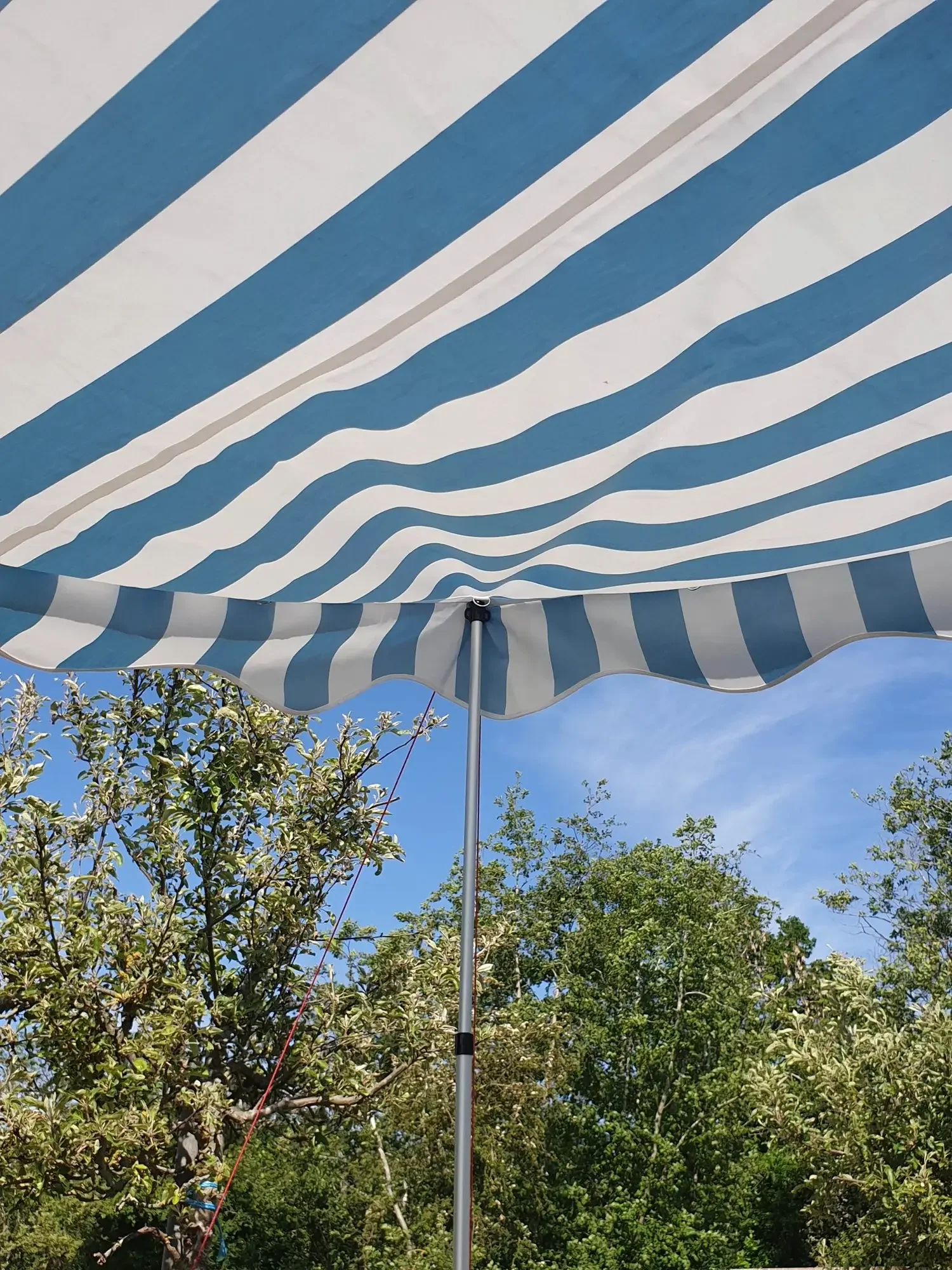 A blue and white striped outdoor patio umbrella with a metal pole, set against a background of green trees and blue sky.