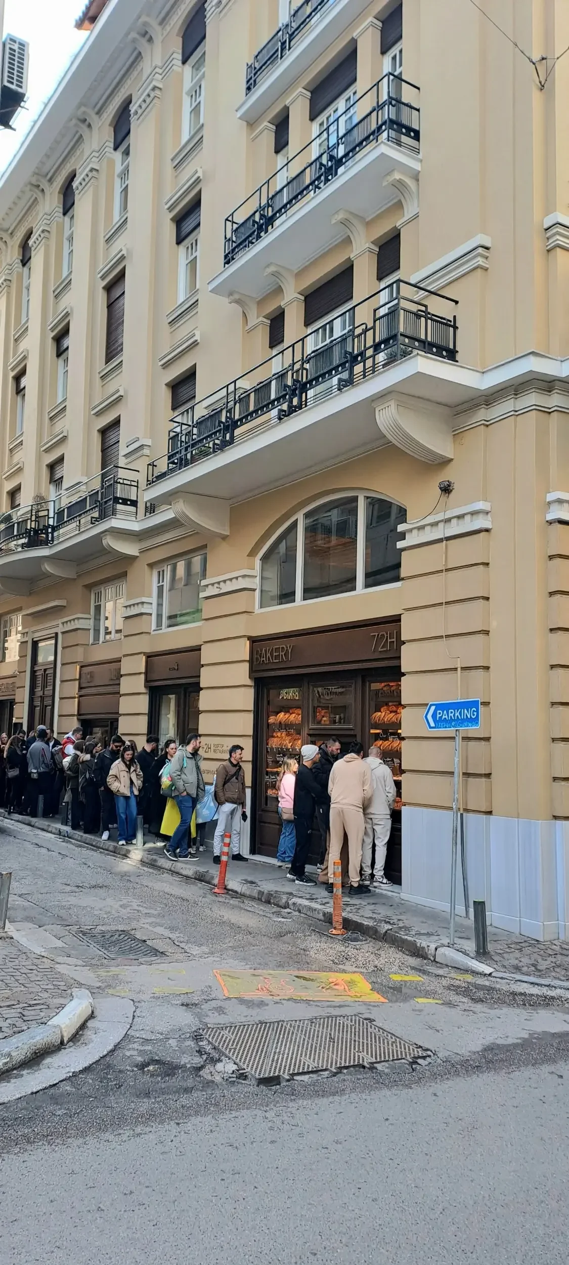 People standing in line outside a bakery storefront on a city street with yellow buildings and balconies, and a blue parking sign.