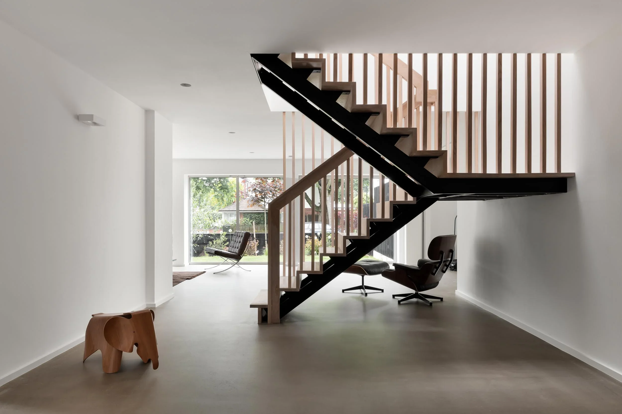 Modern interior with a staircase, large window, and minimal furniture, including a wooden animal-shaped stool and black lounge chairs.