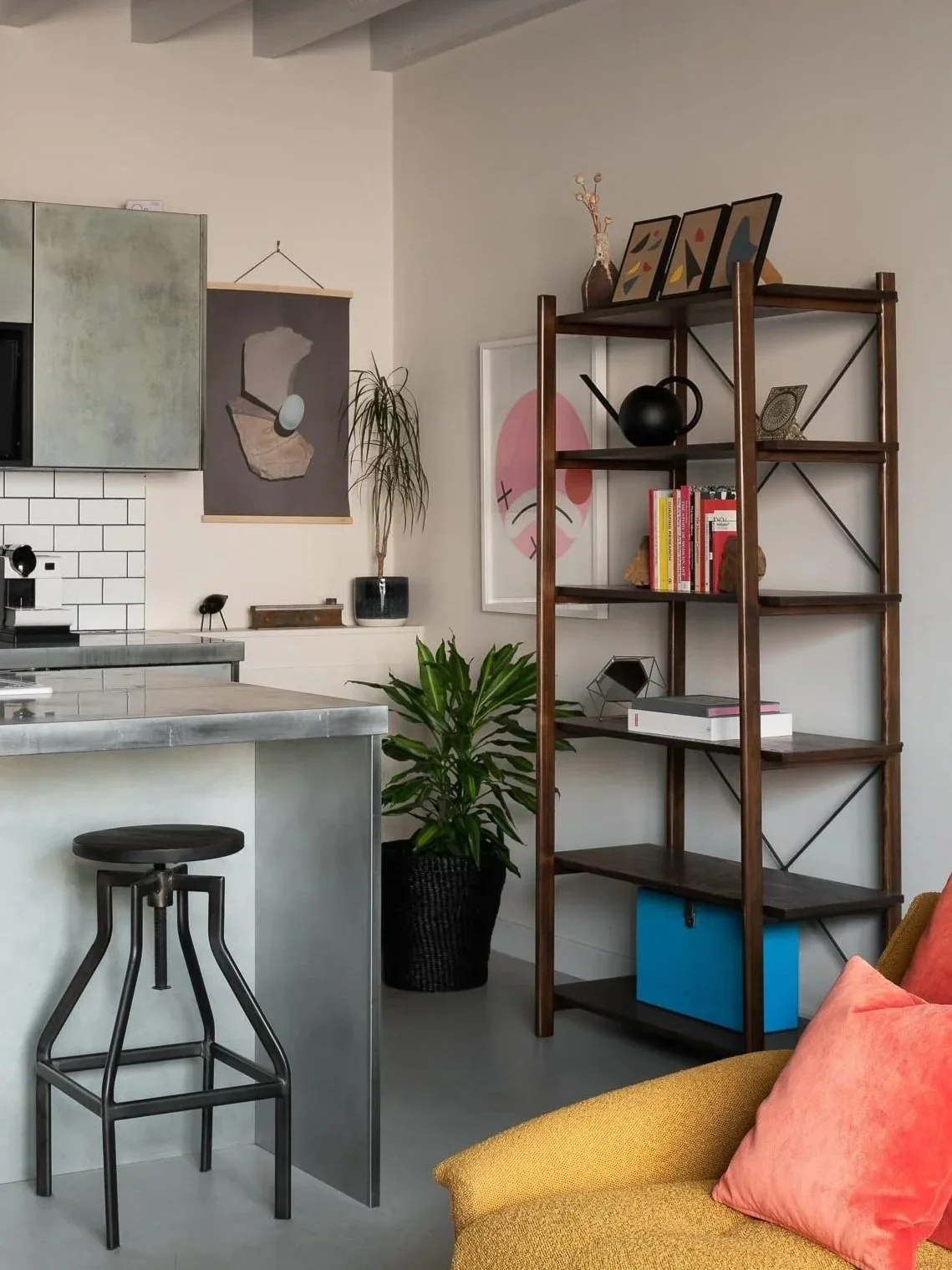 Interior of a modern living space with a yellow sofa and pink cushion, wooden bookshelf with books and decorative items, kitchen counter with a black stool, potted plant, and wall art.