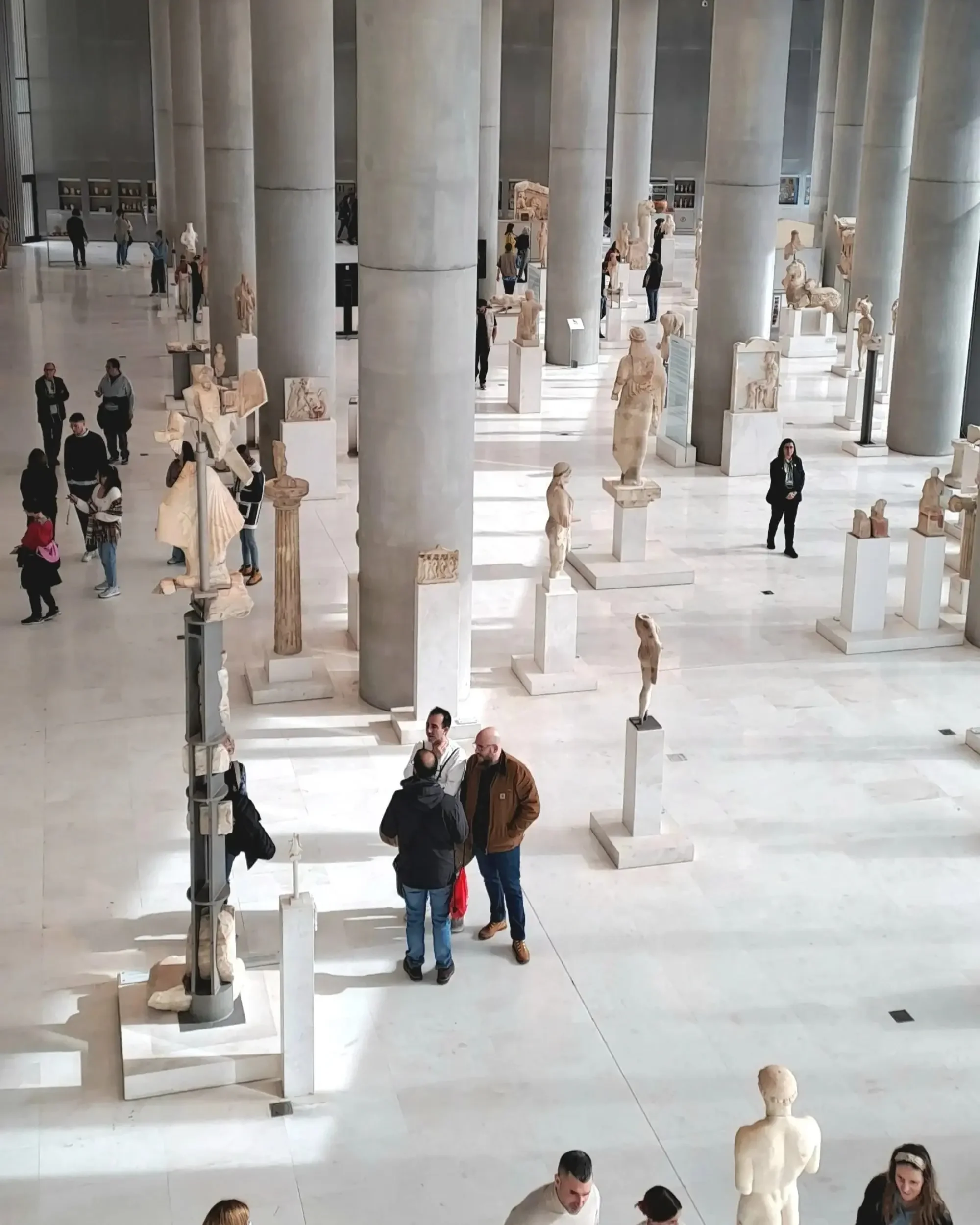 People viewing ancient Greek sculptures and artifacts in a museum hall with large stone columns.