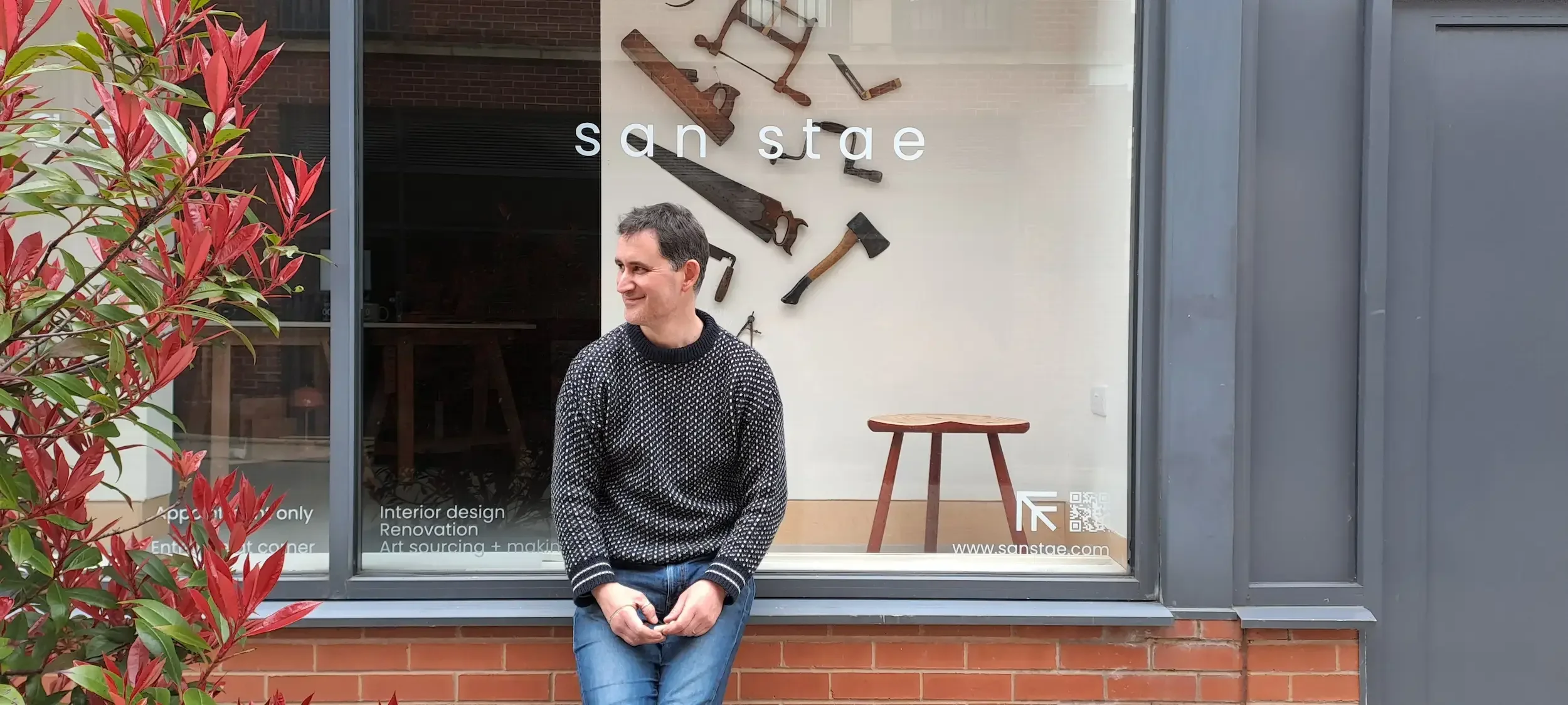 A smiling man in a black and white sweater standing outside a storefront window with tools on the wall inside. Red and green foliage partially blocks the left side of the view.
