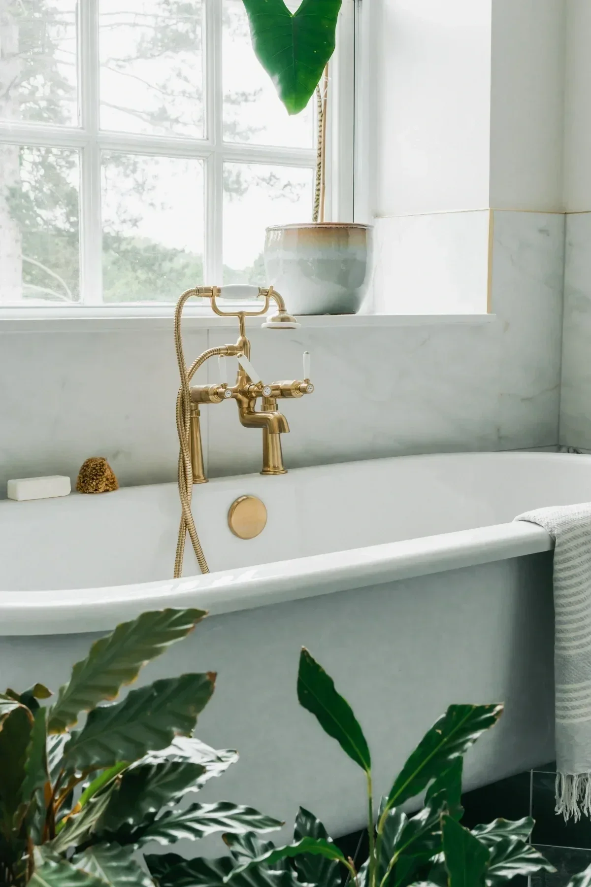 Interior bathroom scene featuring a white clawfoot bathtub with gold fixtures, large window with natural light, a large green leaf plant hanging from the window, a ceramic pot on the windowsill, and a striped towel hanging on the side.