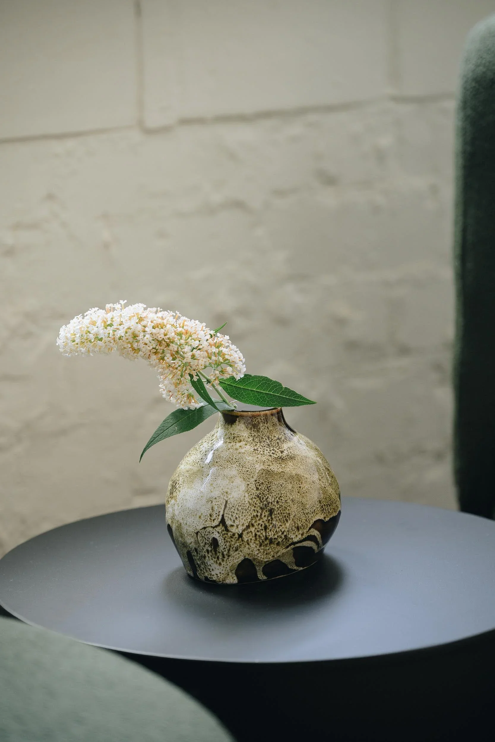 A flower arrangement in a uniquely textured beige and black ceramic vase with a rounded base and narrow neck, placed on a dark round table. The arrangement consists of a single pinkish-white flower with green leaves against a light-colored brick wall background.