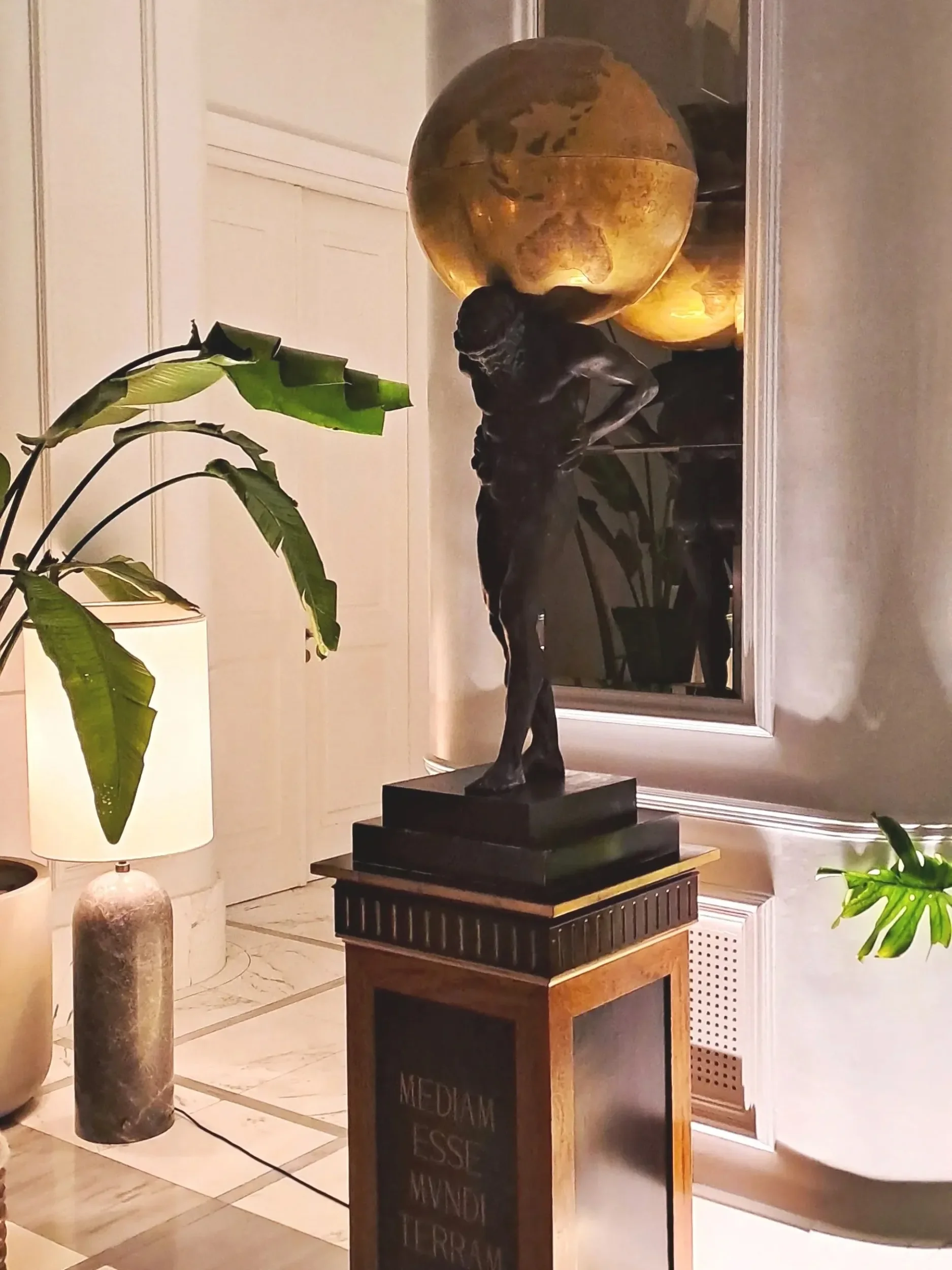 A bronze sculpture of a standing man with a globe on his shoulders, placed on a pedestal near a mirror and houseplants in a warmly lit room.