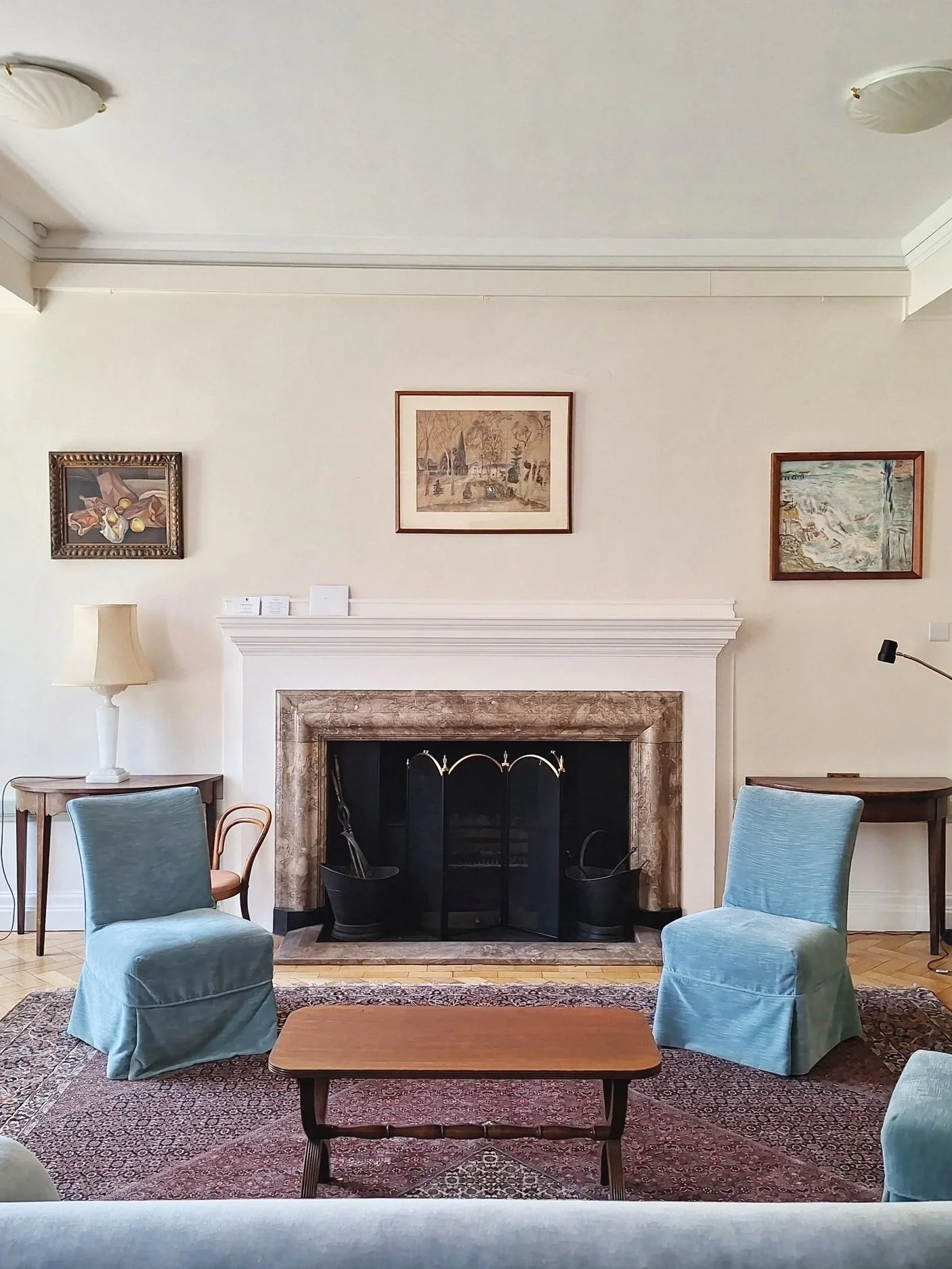 Living room with a fireplace, four blue upholstered chairs, a wooden coffee table, a patterned rug, framed artwork on the wall, a white table lamp, and a small side table.