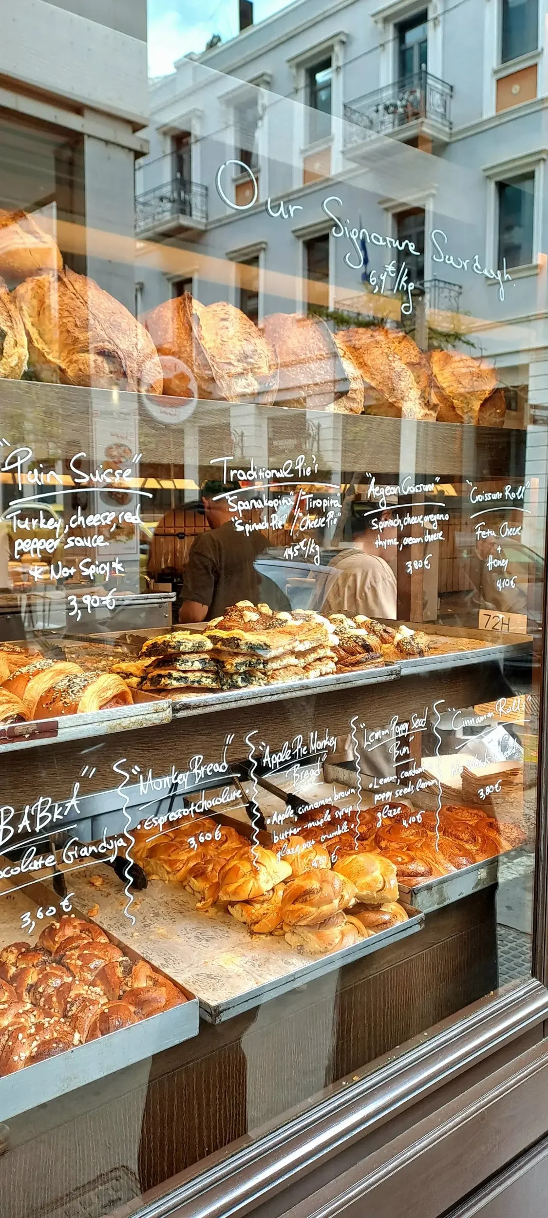 Pastries and baked goods displayed in a bakery window, with handwritten labels for various breads and pies, and city buildings reflected in the glass.