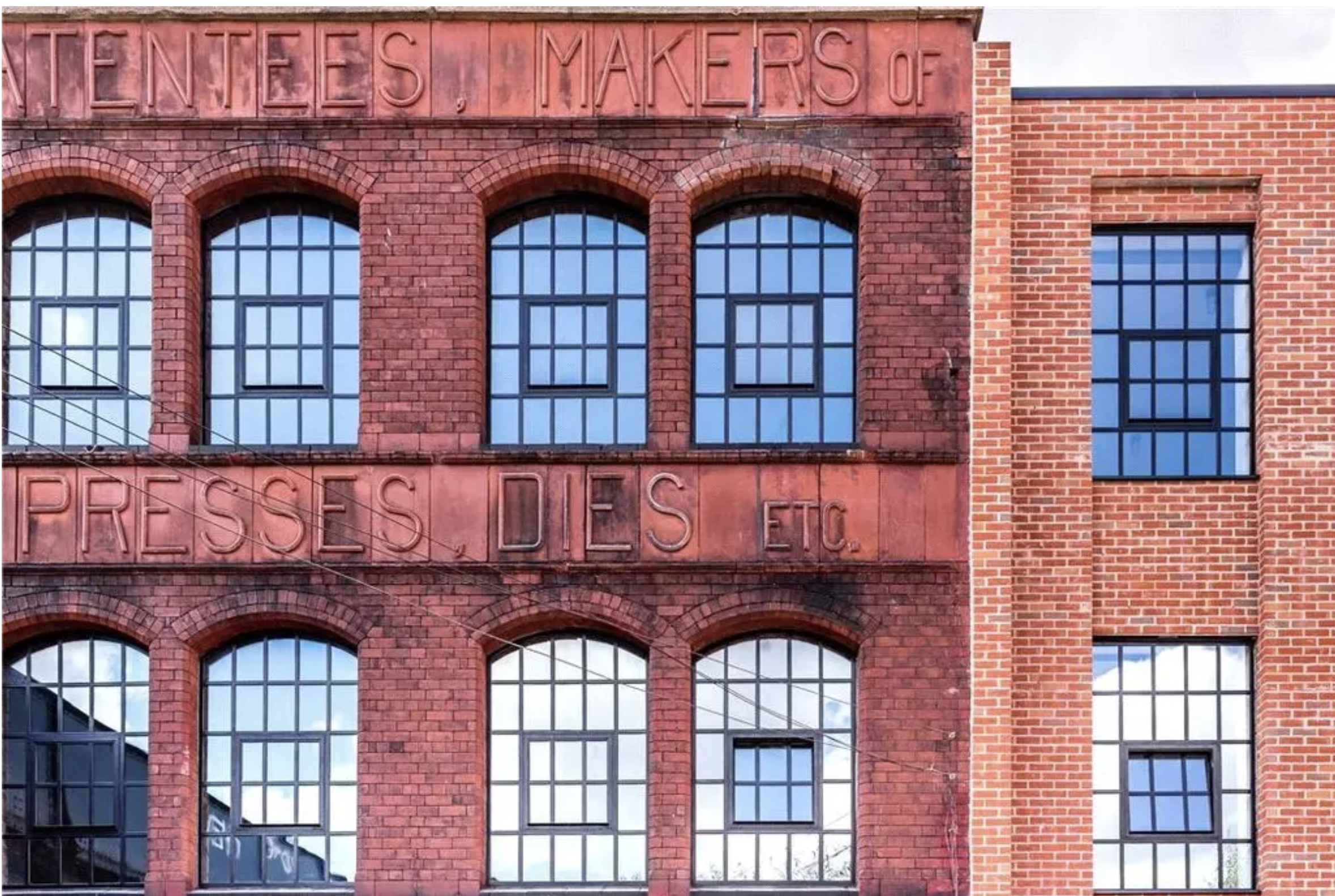 Close-up of a red brick building with large windows, and an embossed sign reading 'TENTEES, MAKERS OF PRESSES, DES ETC.'