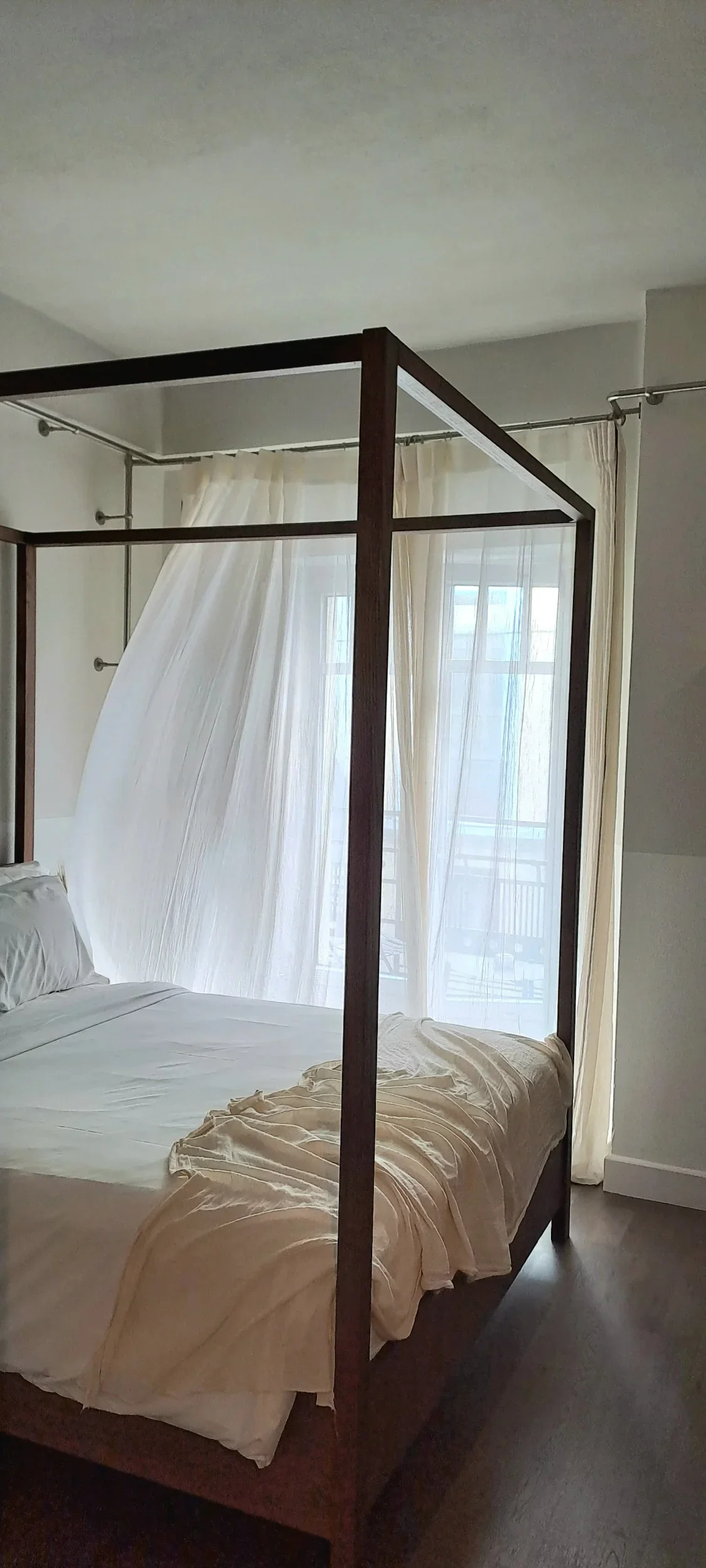 A bedroom with a wooden four-poster bed, white bedding, and sheer curtains on large windows.