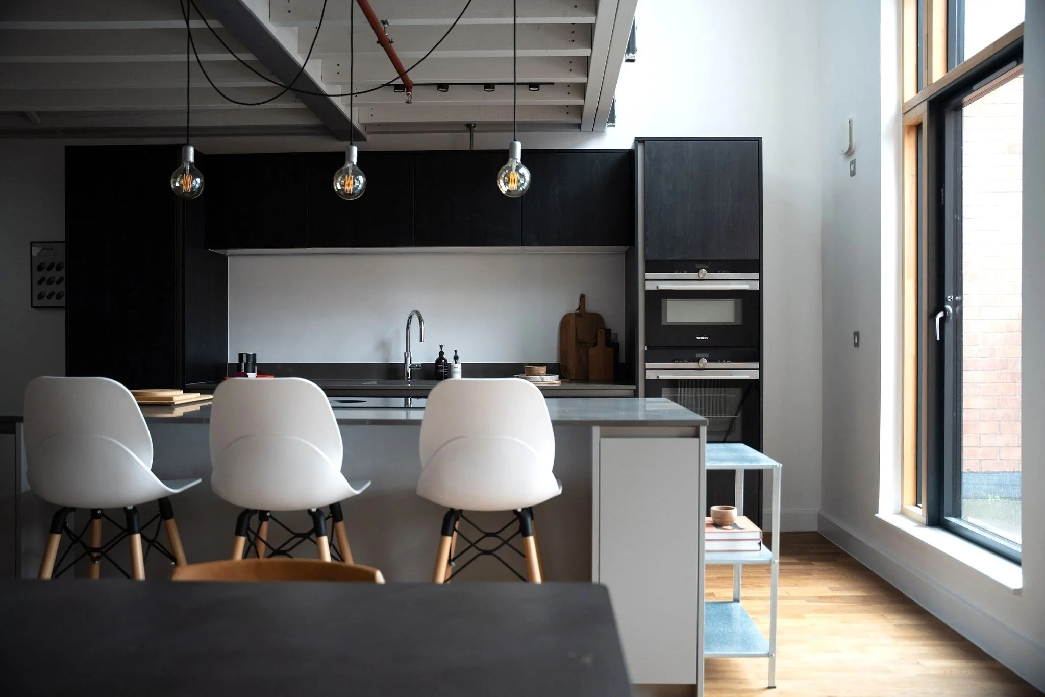 Black wooden kitchen with a kitchen island. Inudstrial looking rafters and 3 industrial pendant lights.