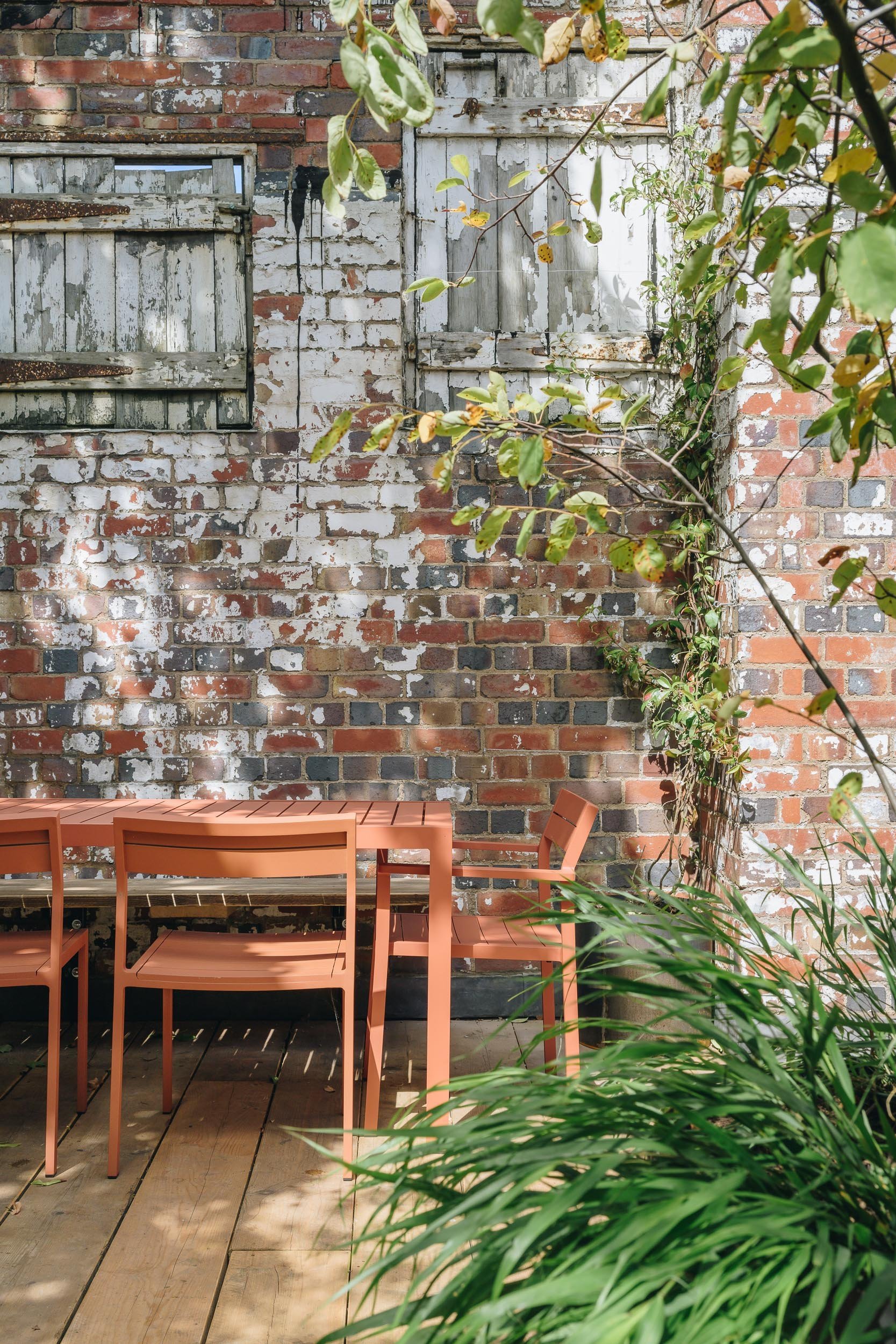 A patio with outdoor furniture including a peach-colored table and chairs, a brick wall with two windows covered with weathered wooden shutters, and green foliage including leafy plants in the foreground.