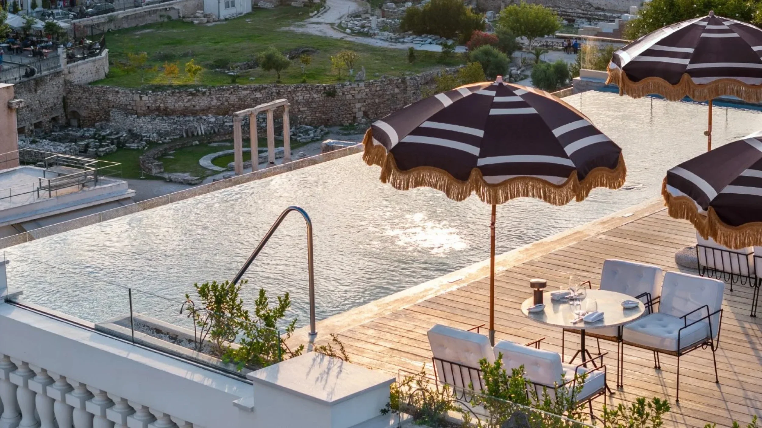 A rooftop pool area with black and white striped umbrellas, outdoor dining tables with chairs, and a view of ancient ruins and greenery in the background.