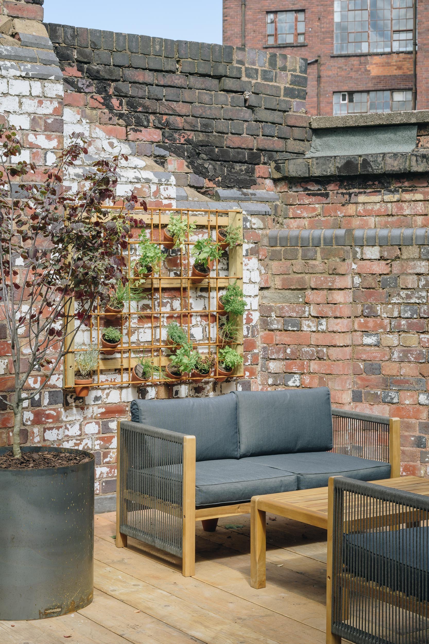 Outdoor patio area with wooden flooring, a blue cushioned outdoor sofa, a matching armchair, a wooden coffee table, potted plants, and a large plant in a gray pot. The backdrop features a brick wall with a mounted wooden plant holder and an apartment building in the distance.