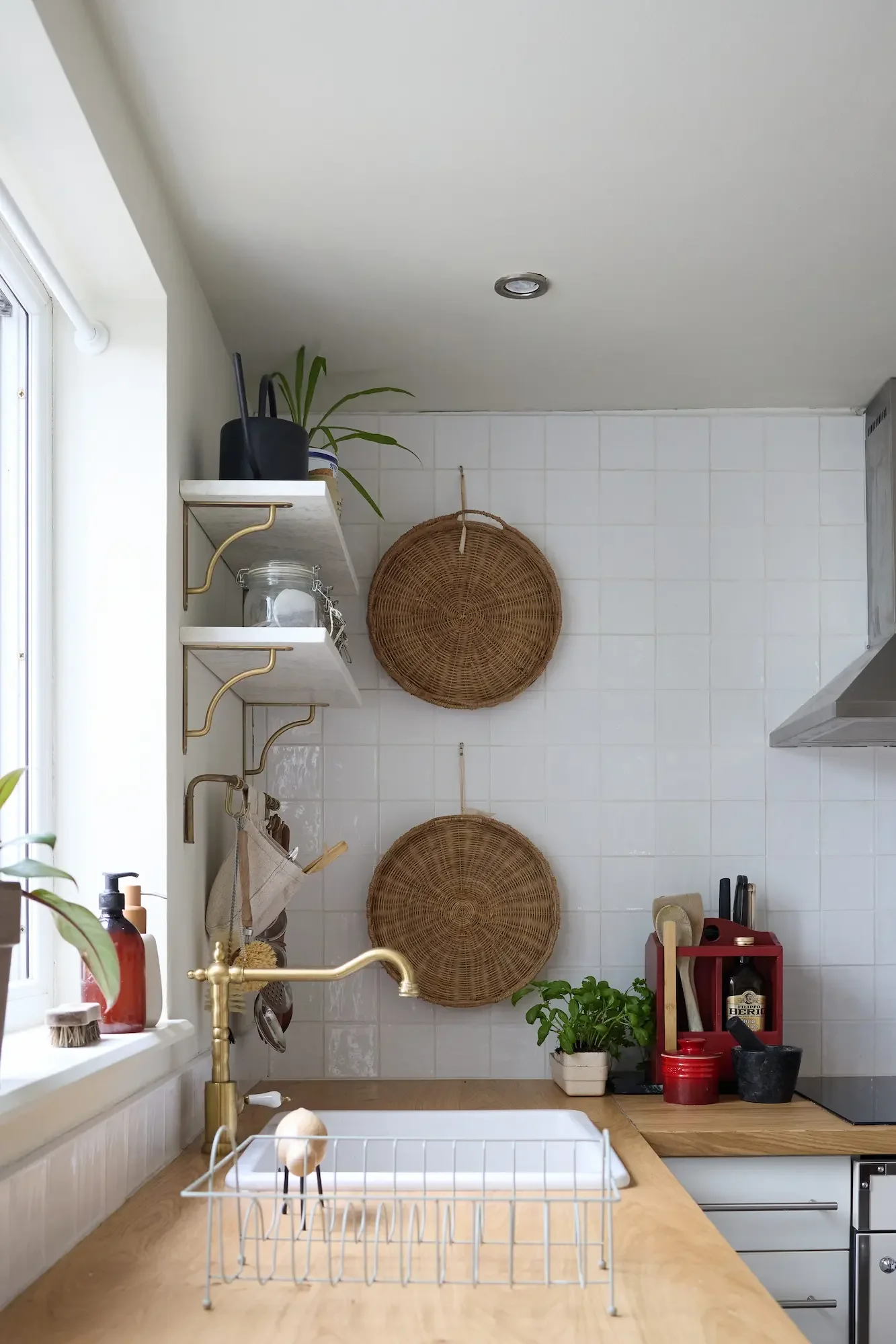 A kitchen with white walls, a wooden counter, hanging woven baskets, a small plant, shelves with various items, a sink with a gold faucet, and a window letting in natural light.