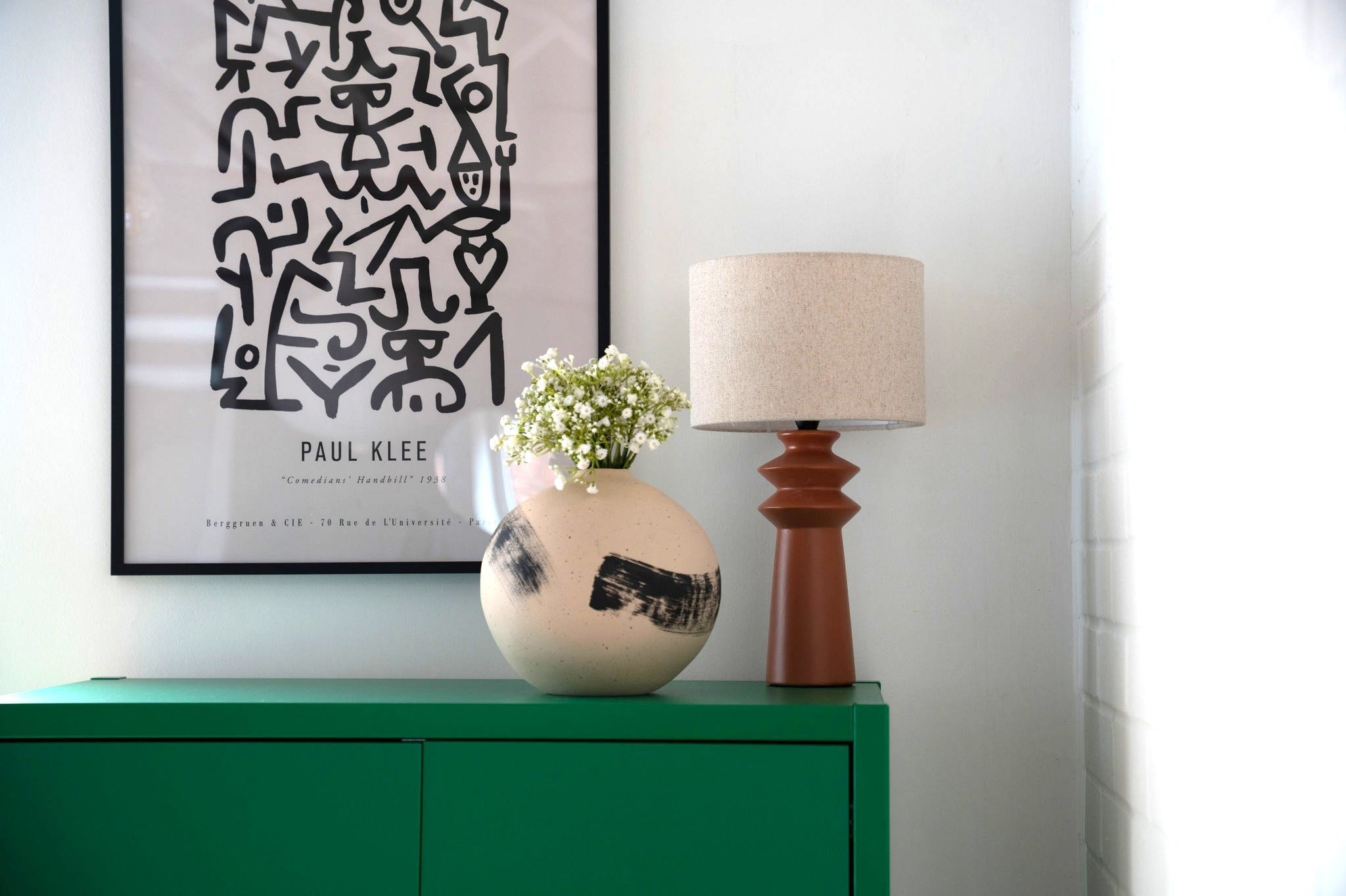 Ceramic vase with a paint stripe. Brown base table lamp sat on a green cupboard. Paul Klee print in a black frame. Design by San Stae Interior Design and Art Studio.