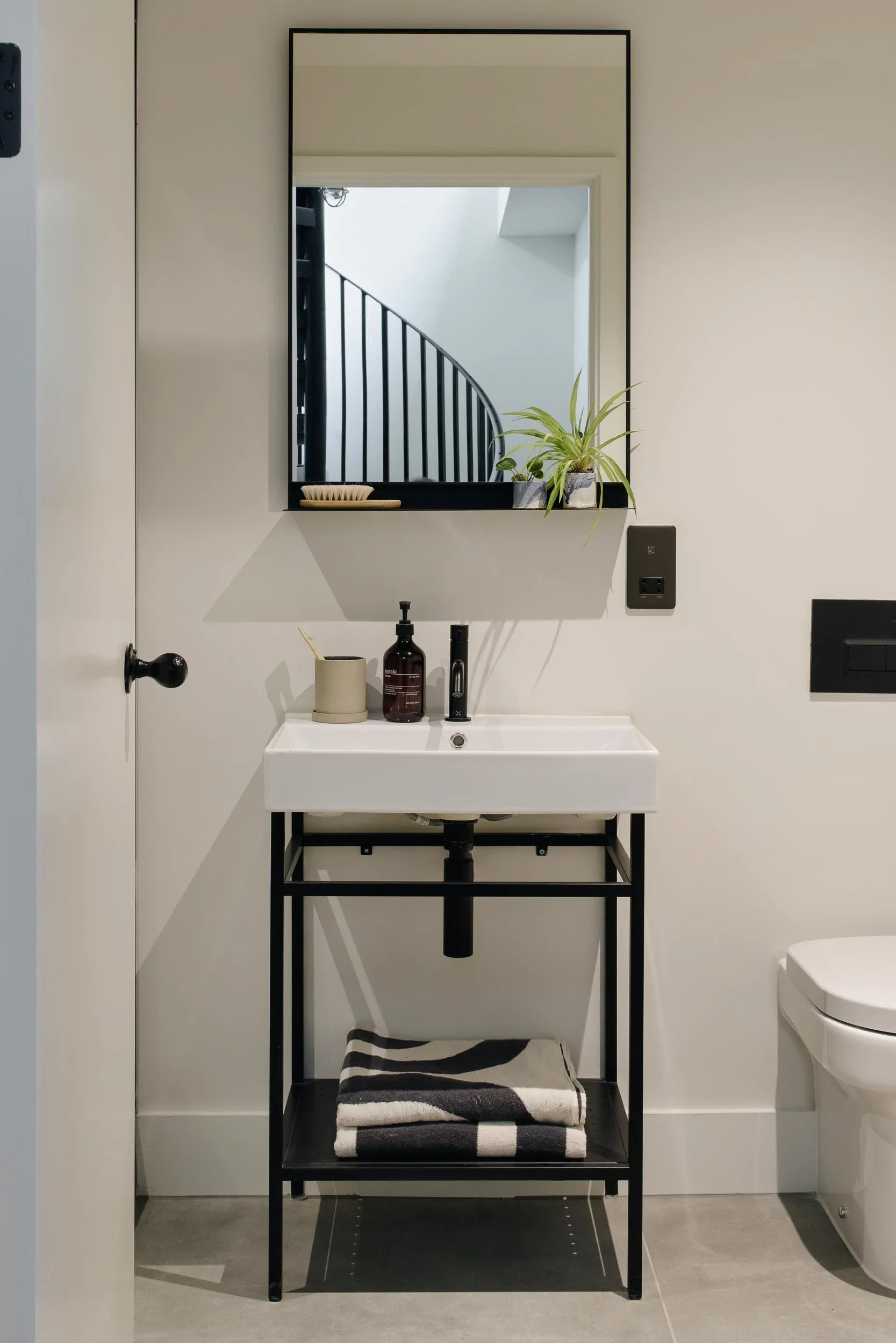 Minimalist bathroom with white sink on black metal stand, black and white striped towel, mirror reflecting staircase, small plant on the shelf, soap dispenser, toothbrush holder, and a soap container.
