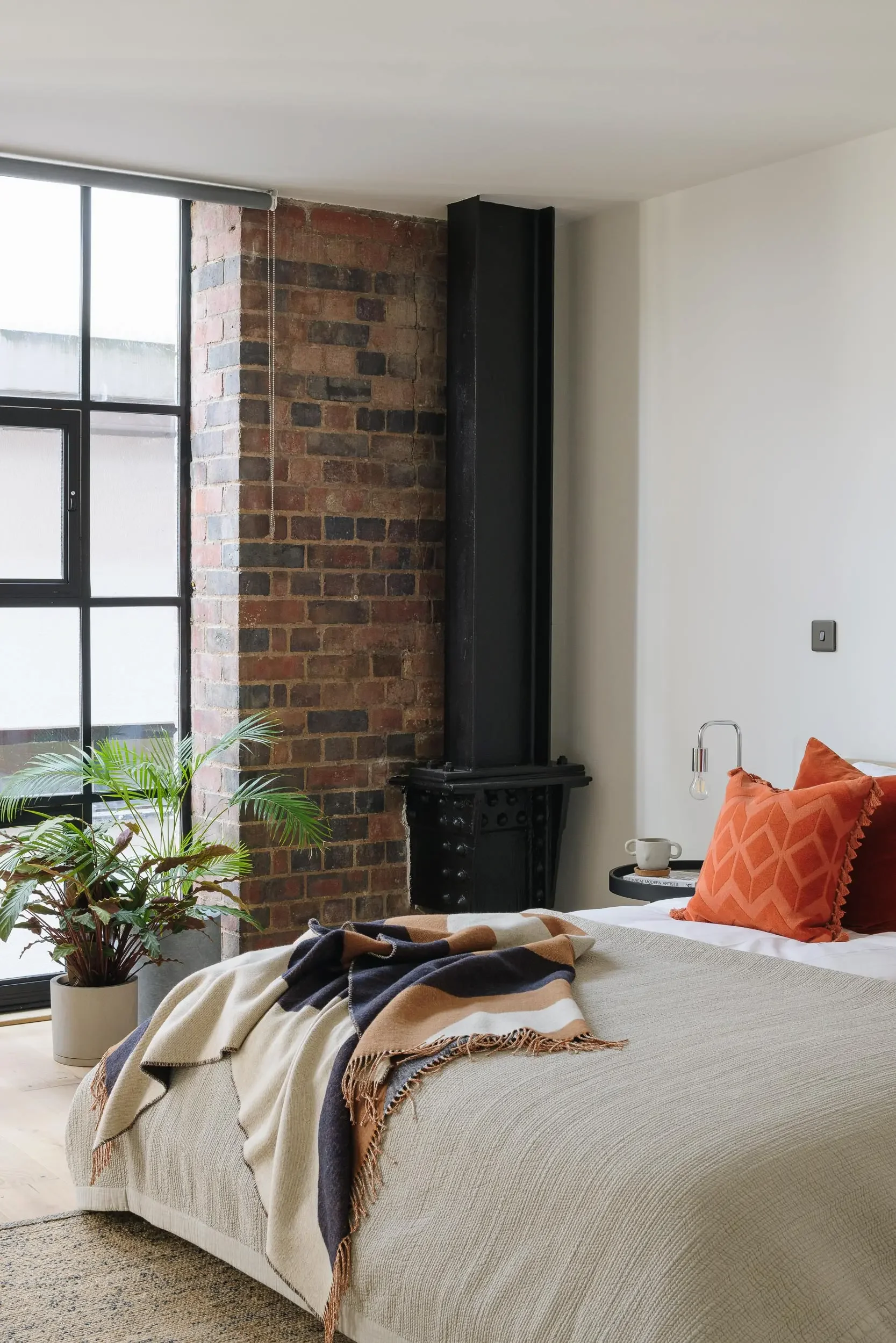 A cozy bedroom corner with a bed covered in a beige textured blanket, topped with an orange geometric pillow, and a plaid throw blanket. A potted plant is placed on the floor next to the bed. In the background, there is a black fireplace, a large window with black frames, a brick wall, and a white wall with a small electrical outlet.