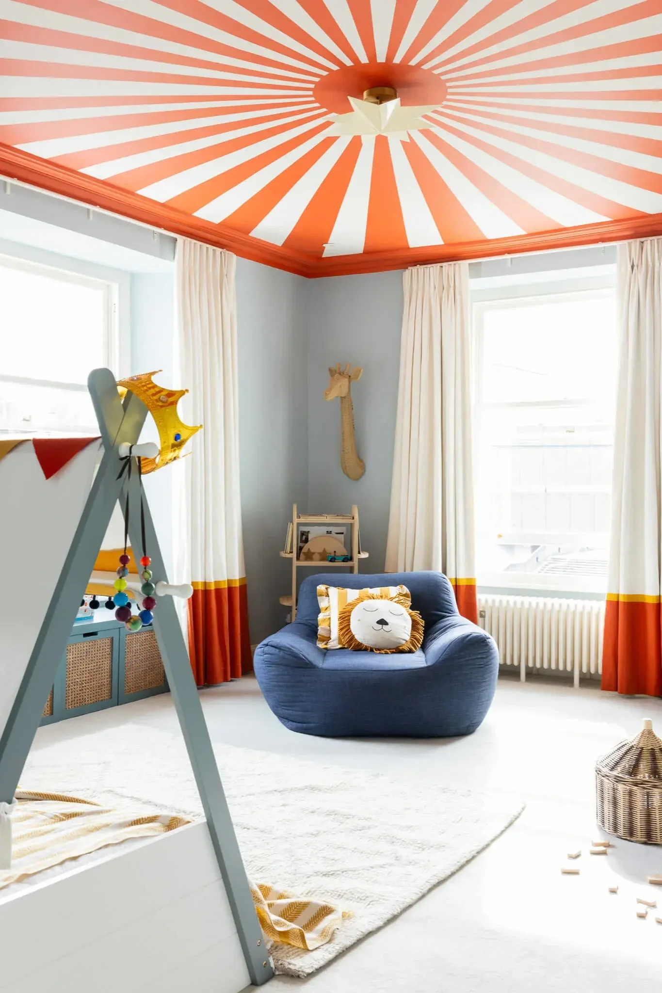 A cozy children’s room with white walls and large windows with white curtains. The ceiling has a bright orange and white striped pattern with a star-shaped light fixture. There is a small plush lion pillow on a blue armchair, a wooden giraffe wall decoration, and an easel with a colorful crown and beads hanging from it. A soft white rug covers the floor, and a wicker basket is in the corner.