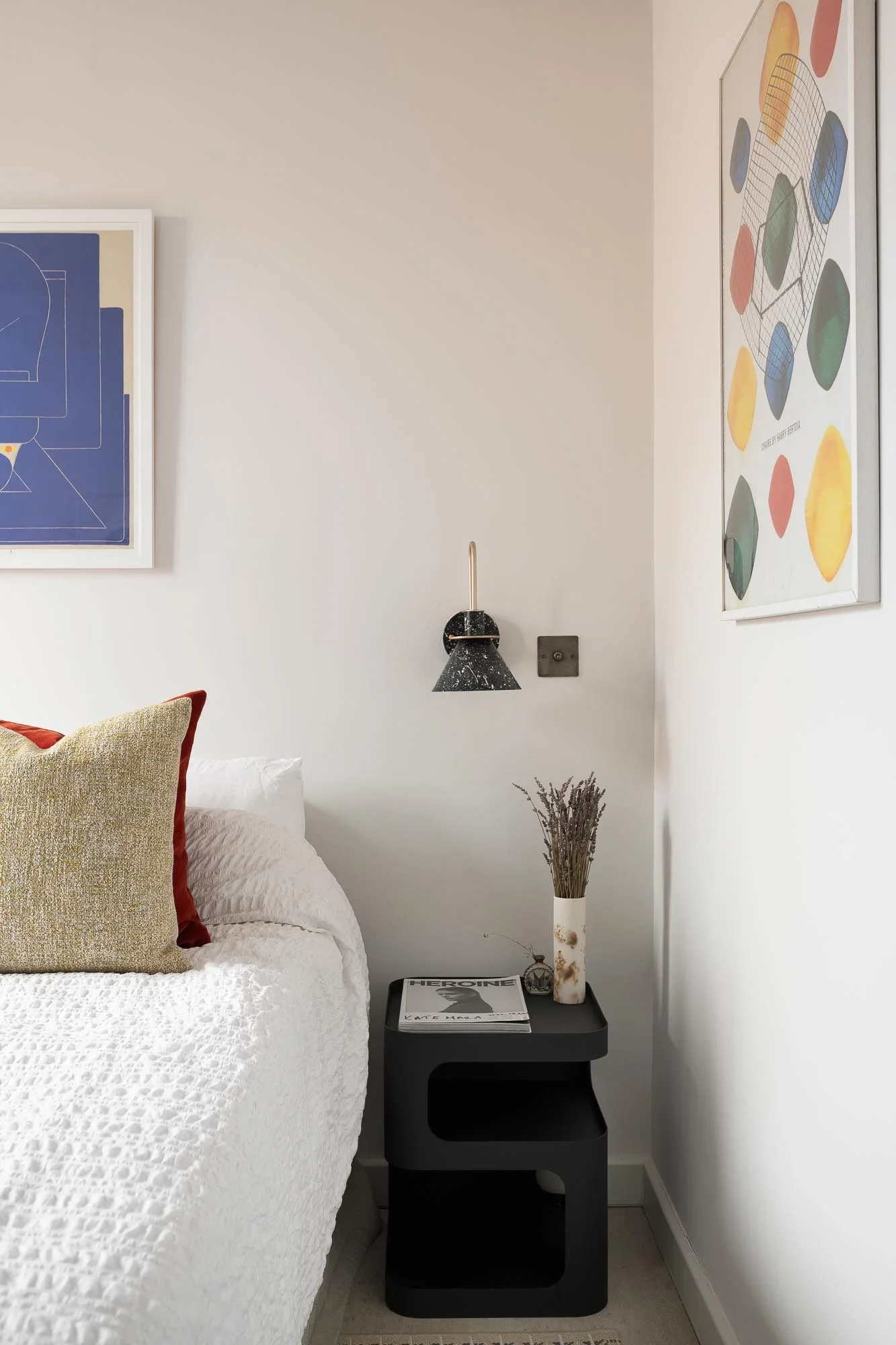 A bedroom corner with white walls, a bed with white bedding and beige and red pillows, a black side table holding a magazine titled "HEROIN" and a decorative vase with dried plant stems. Above the bed, there's a black wall-mounted lamp and two colorful framed artworks on the wall.