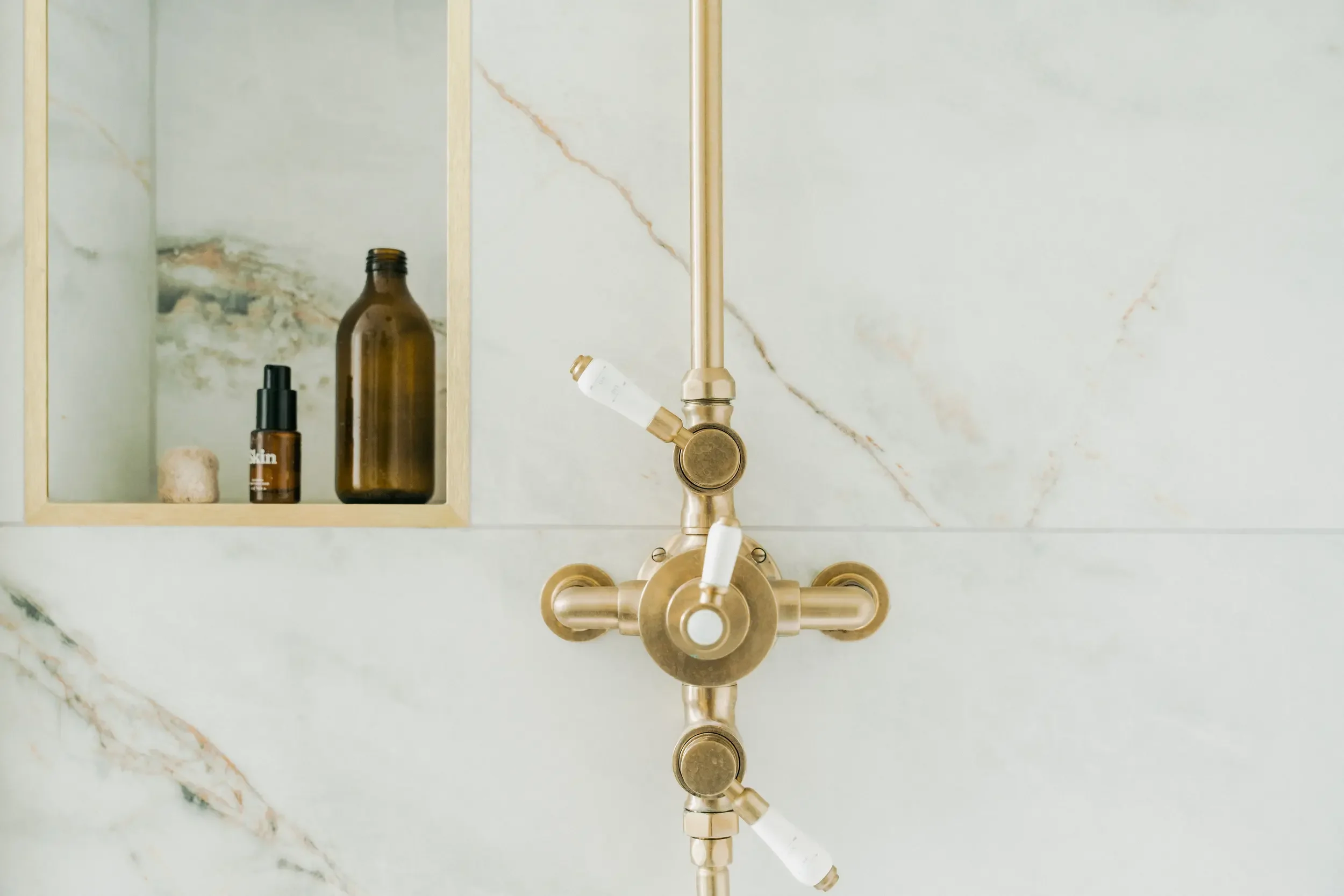 Close-up of a gold-colored shower valve with four white knobs, set into a marble wall with a built-in niche containing brown bottles and a small rock.