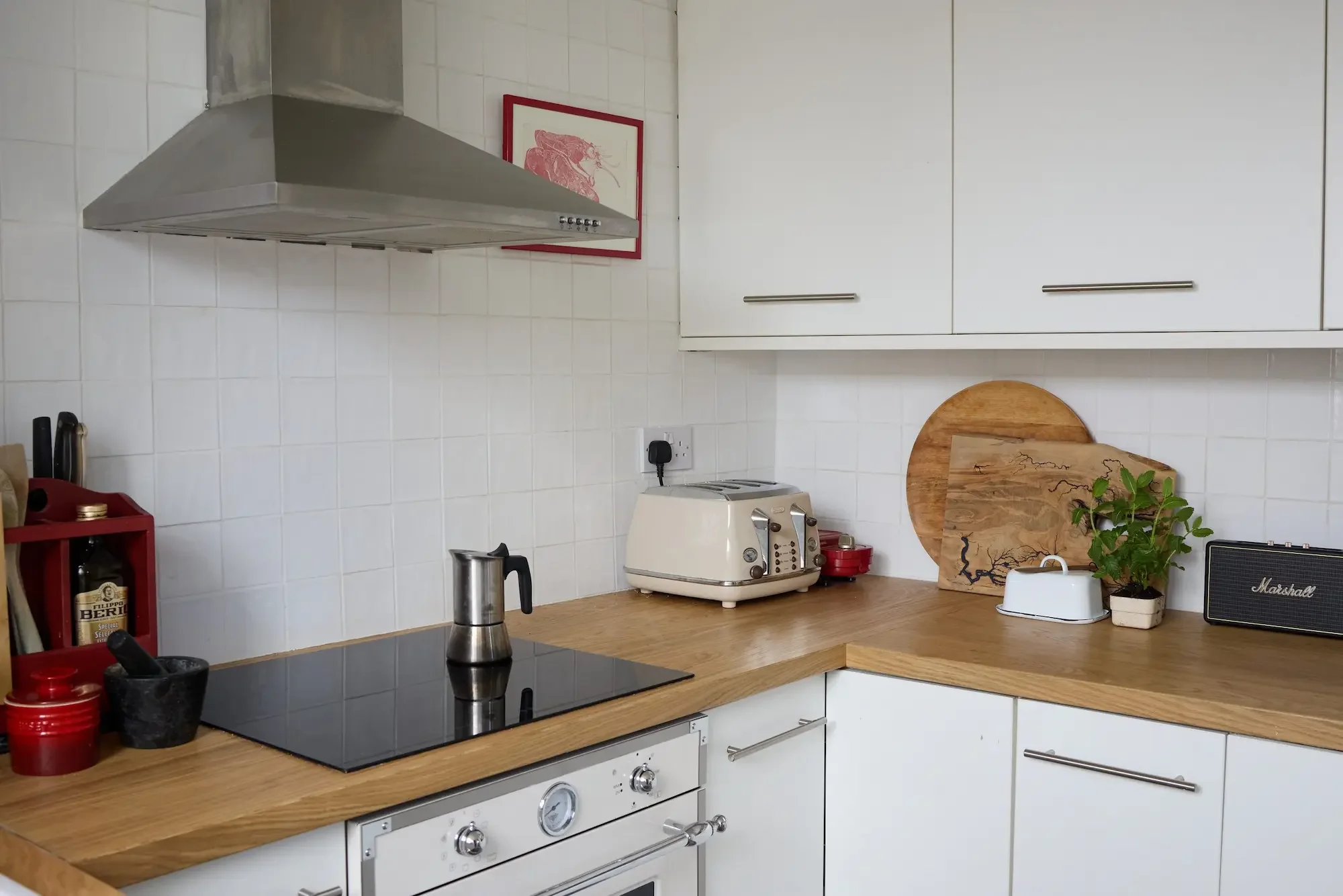 A kitchen with white tile walls, wooden countertops, and white cabinets. There is a stainless steel range hood, a framed picture of a lobster, a toaster, a cutting board with a plant, and various small kitchen items such as a red spice jar, a black mortar and pestle, and a bottle of Italian hot sauce on the countertop.