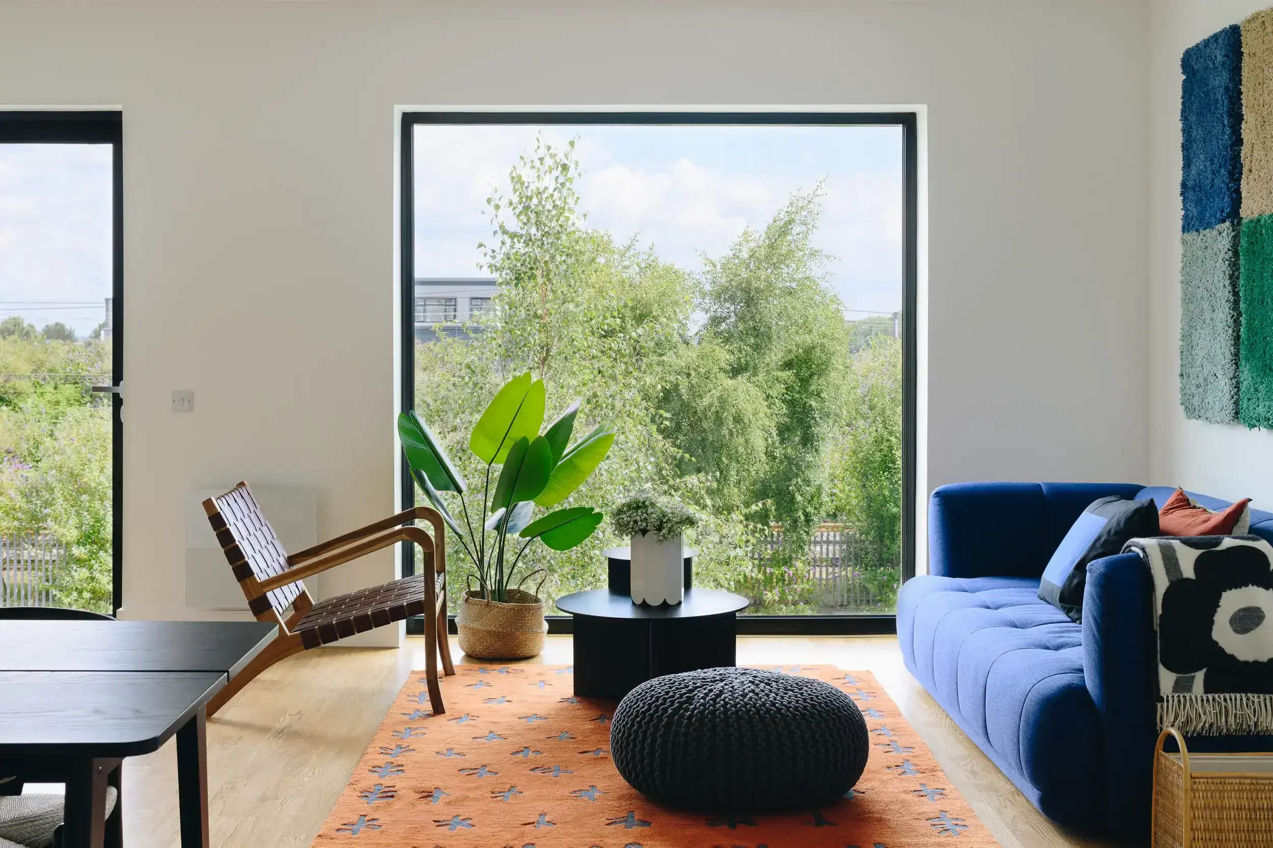 Bright living room with large window overlooking green trees, featuring a blue sofa, a black round coffee table, a woven pouf, a wooden chair, potted plants, and colorful wall art.