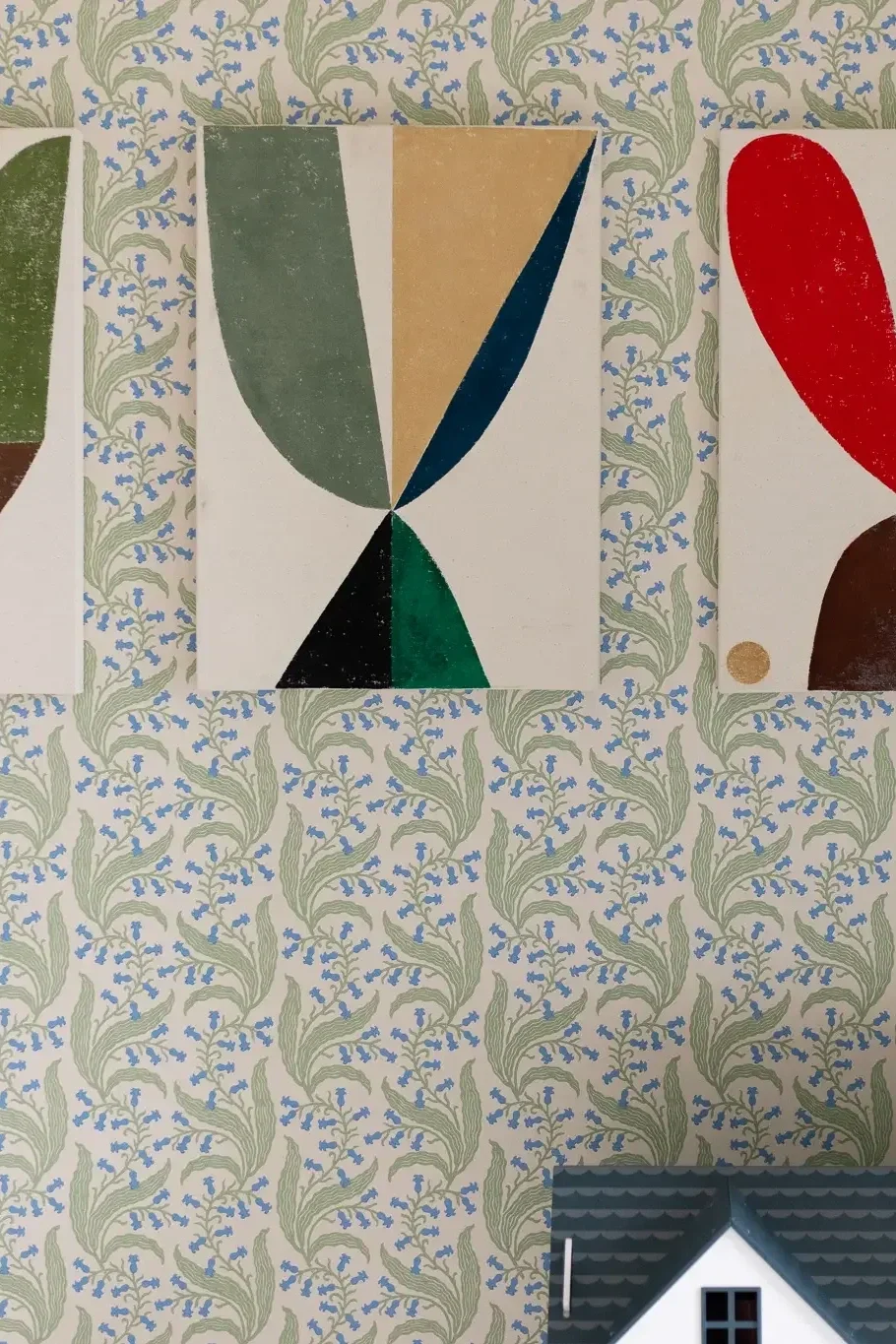 Close-up of wall with decorative wallpaper featuring beige, blue, and green floral pattern, showing portions of abstract artwork and a small part of a model house roof at the bottom.