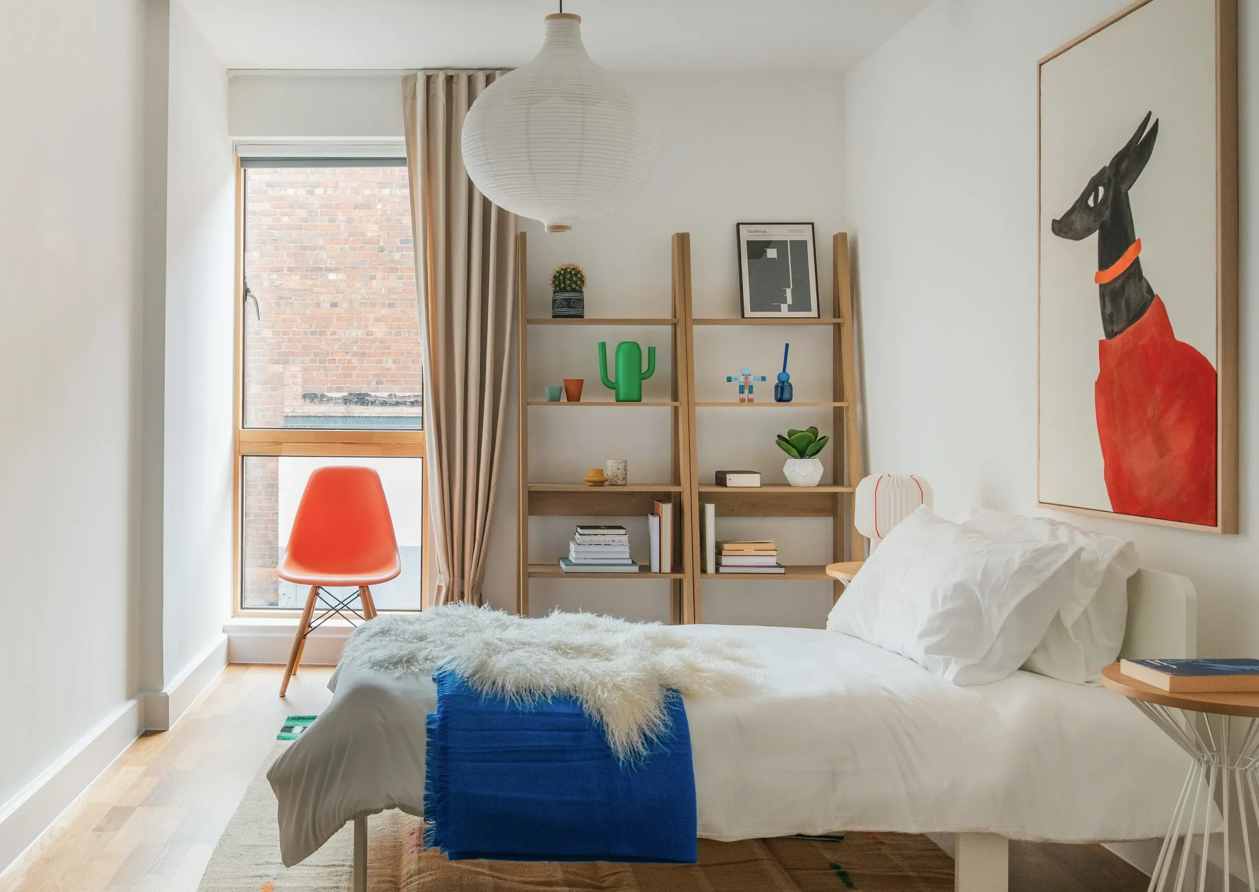 A bedroom with a white bed, a wooden bookshelf with plants and framed artwork, a bright window with beige curtains, a orange chair, and a paper lantern hanging from the ceiling.