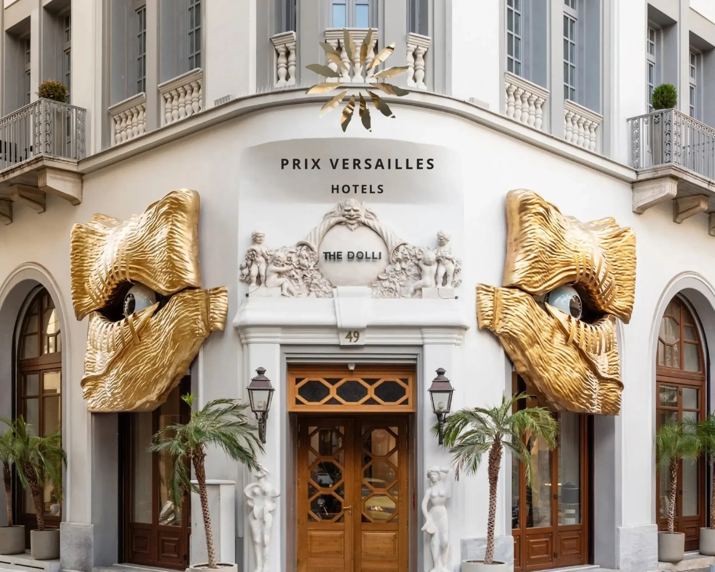 Exterior of a hotel with large gold sculptures of eyes on either side of the entrance, palm trees, and classical architectural details.