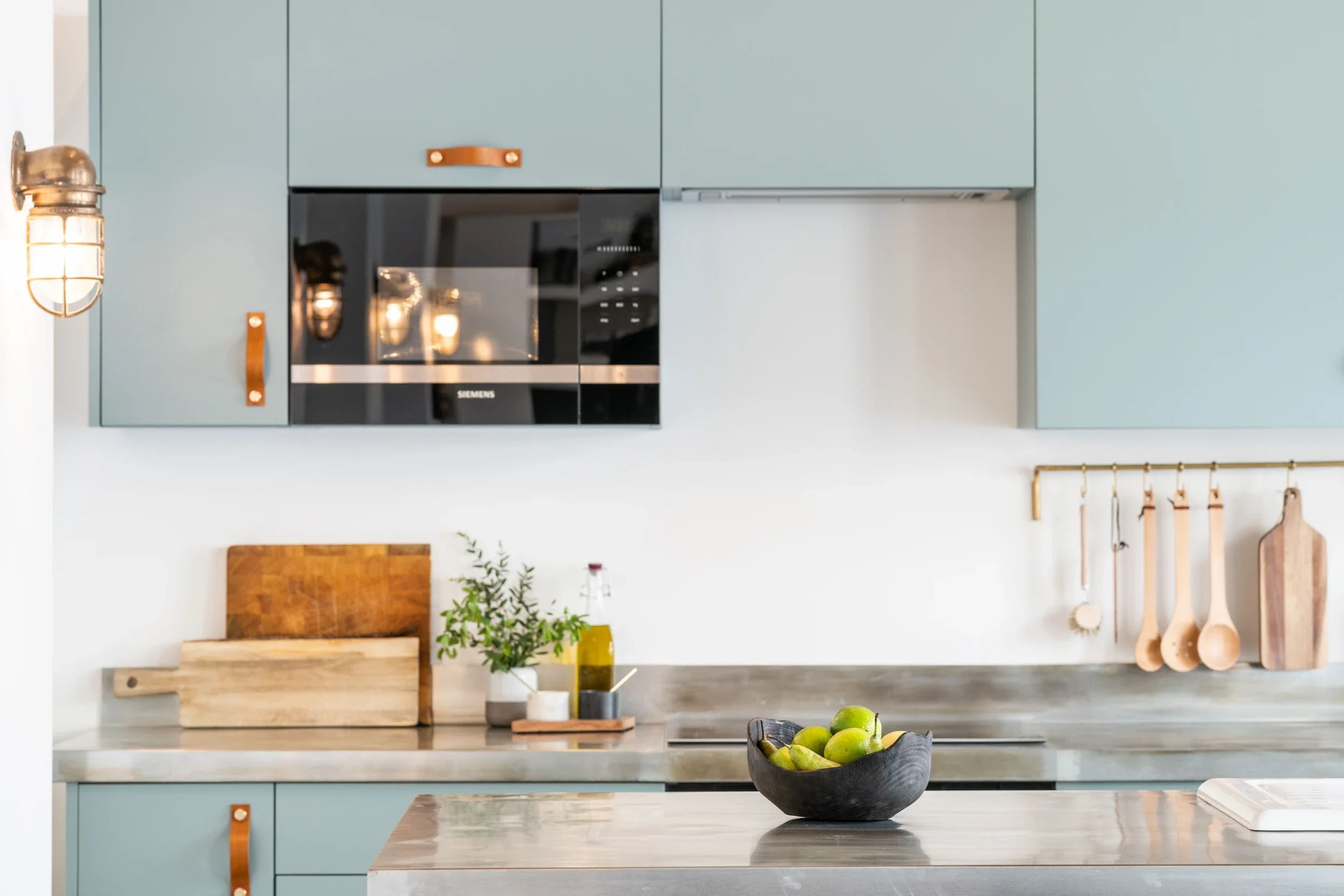 A modern kitchen with light blue cabinets, a black microwave oven, and kitchen utensils hanging on a rack. There is a wooden cutting board, a plant in a yellow pot, and a bowl of green apples on the countertop.