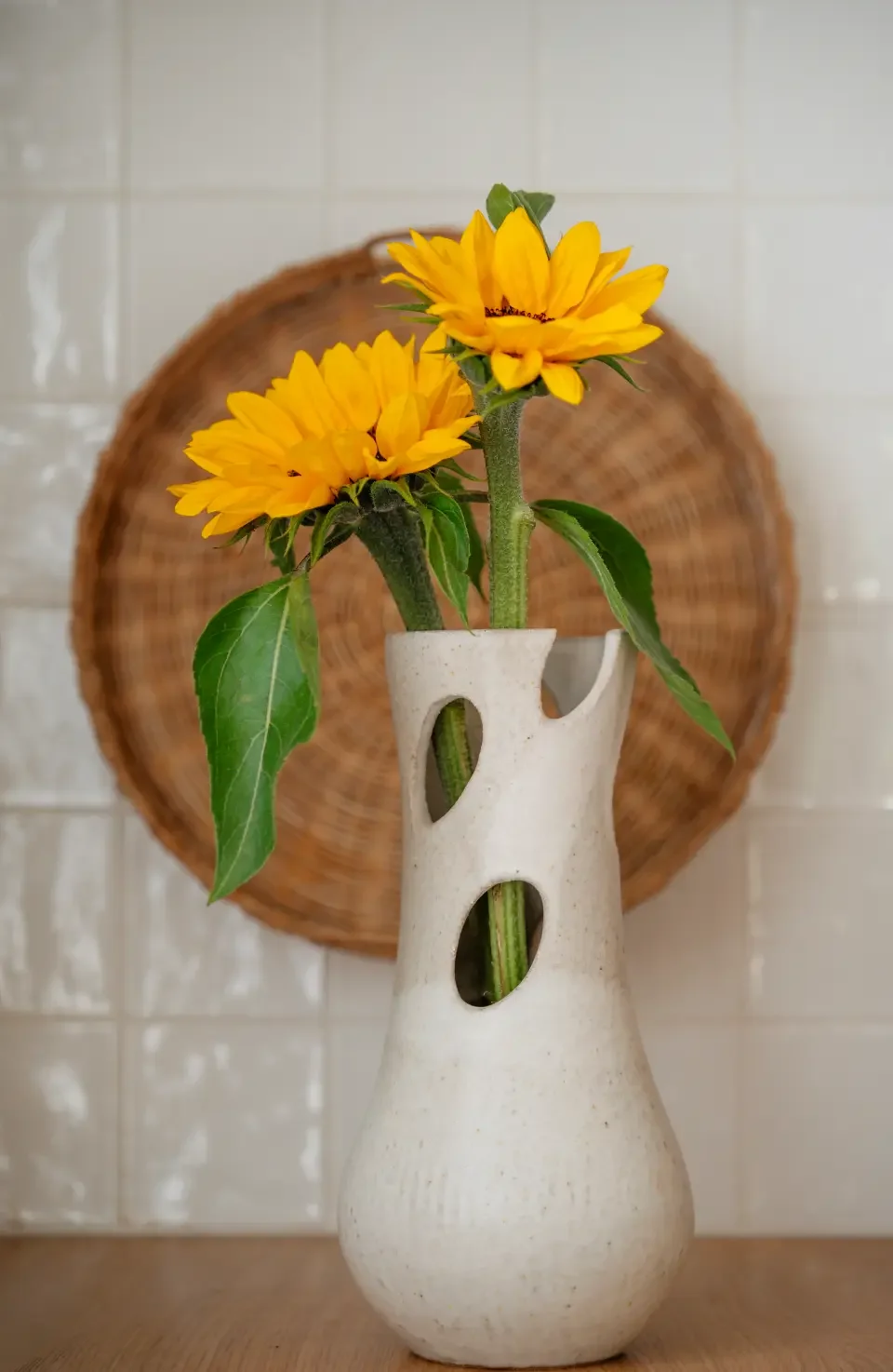 Yellow sunflowers in a white ceramic vase with cutout holes, against a background with a circular wicker basket on a tiled wall.