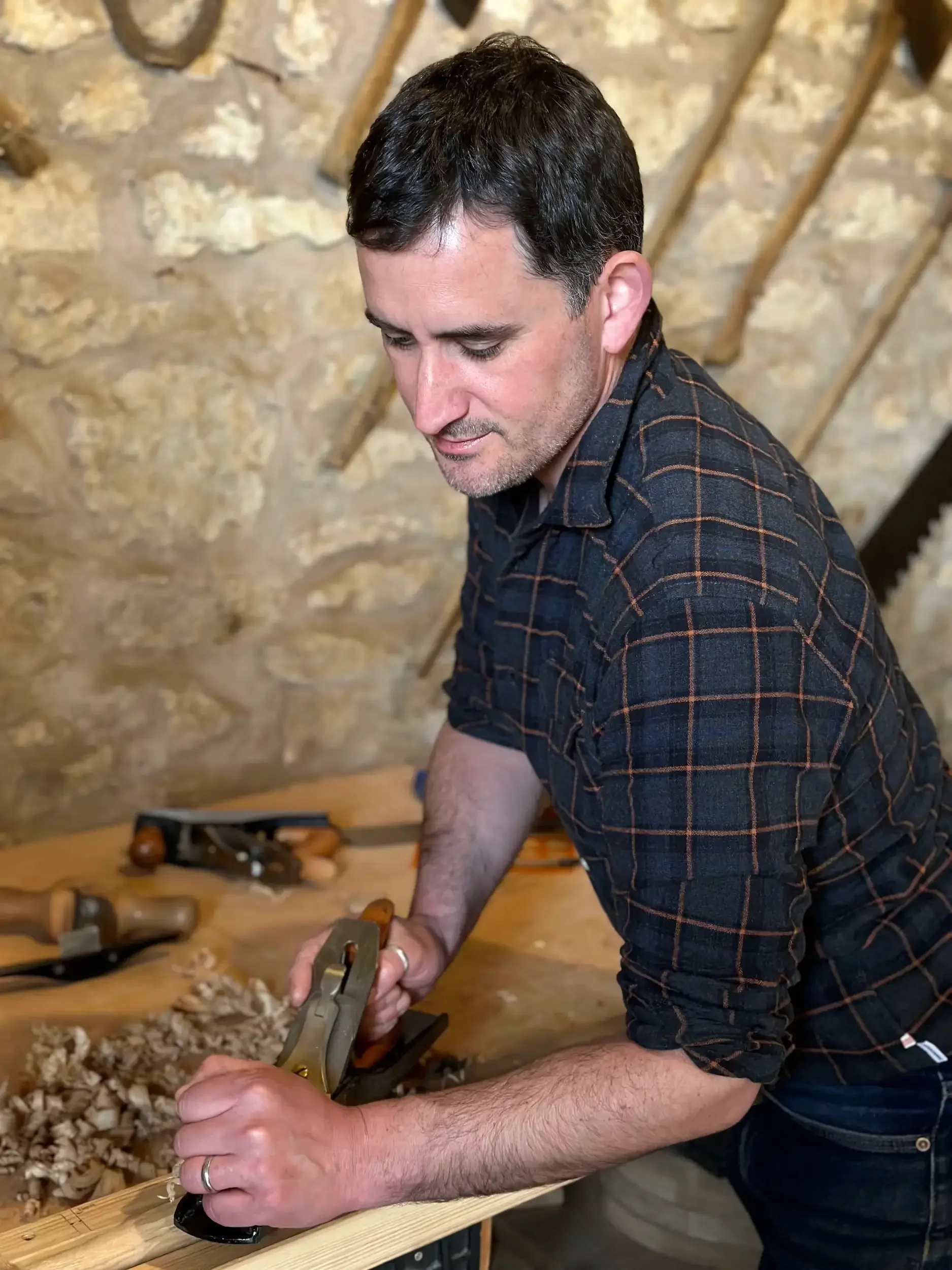A man with dark hair, wearing a checkered shirt, working with a hand plane on a wooden piece in a woodworking shop.