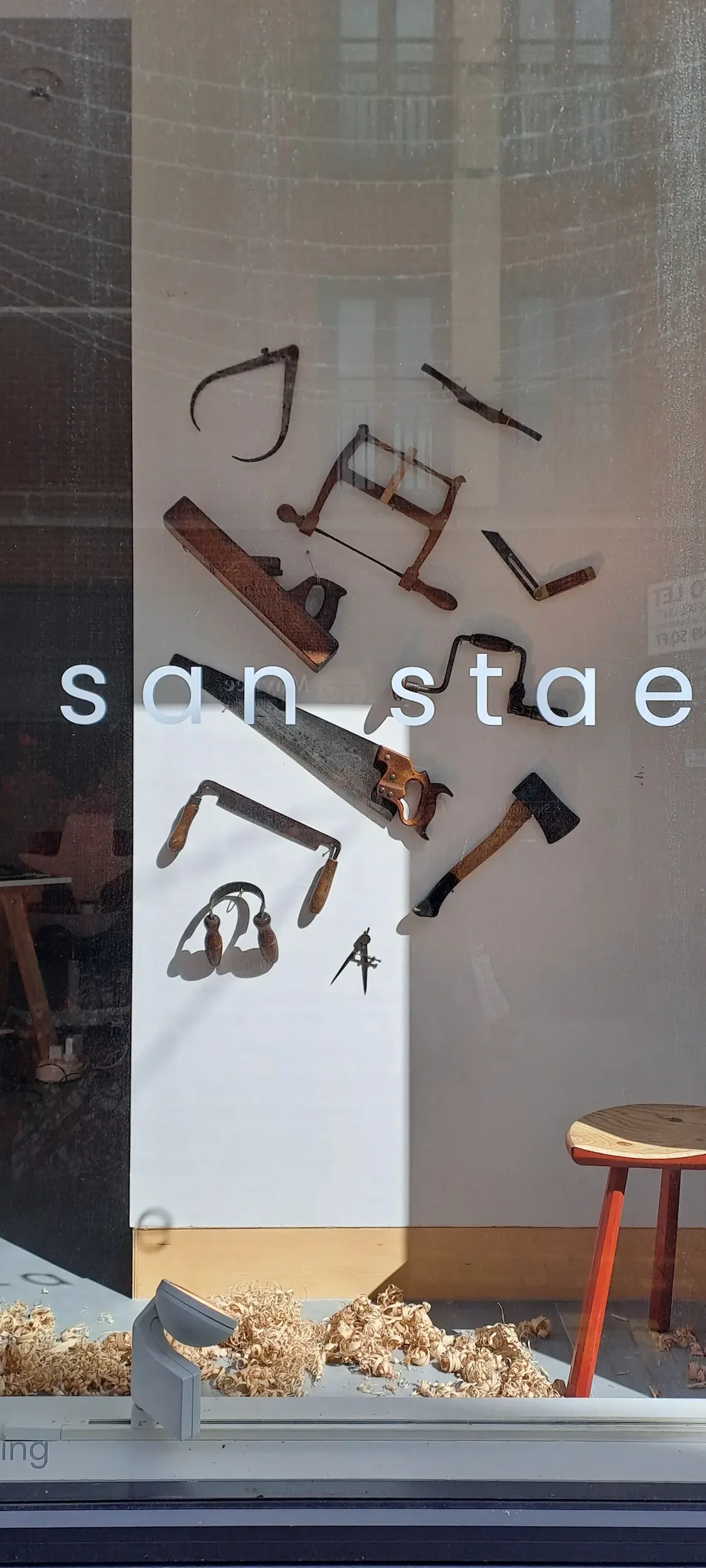 Kitchen tools like a hand saw, a hammer, a wrench, and other tools displayed on a window with the words 'san stade' and a bunch of wood shavings below.